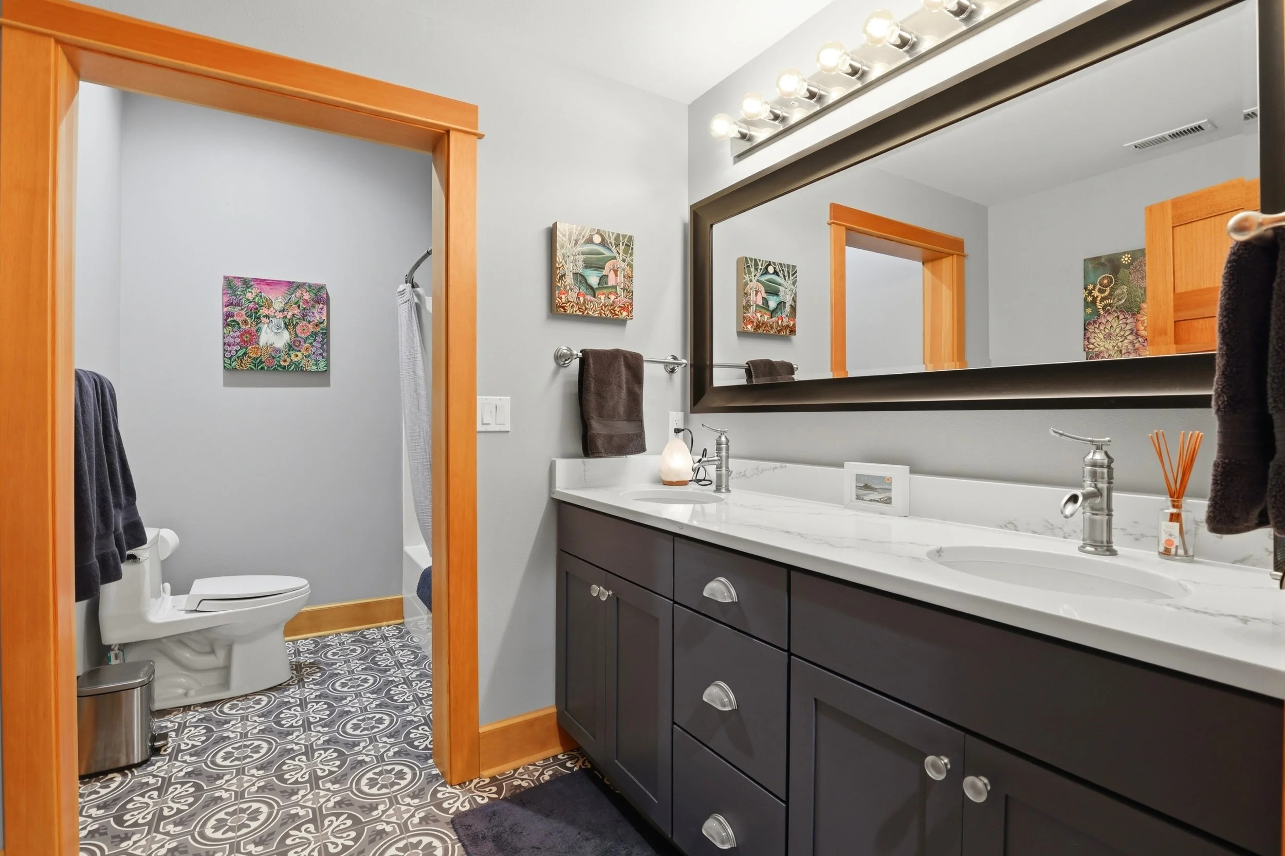 A modern bathroom with a white countertop vanity with black cabinetry, a large mirror, and decorative light fixtures. There's a toilet with artwork above, wooden door frames, and patterned floor tiles.