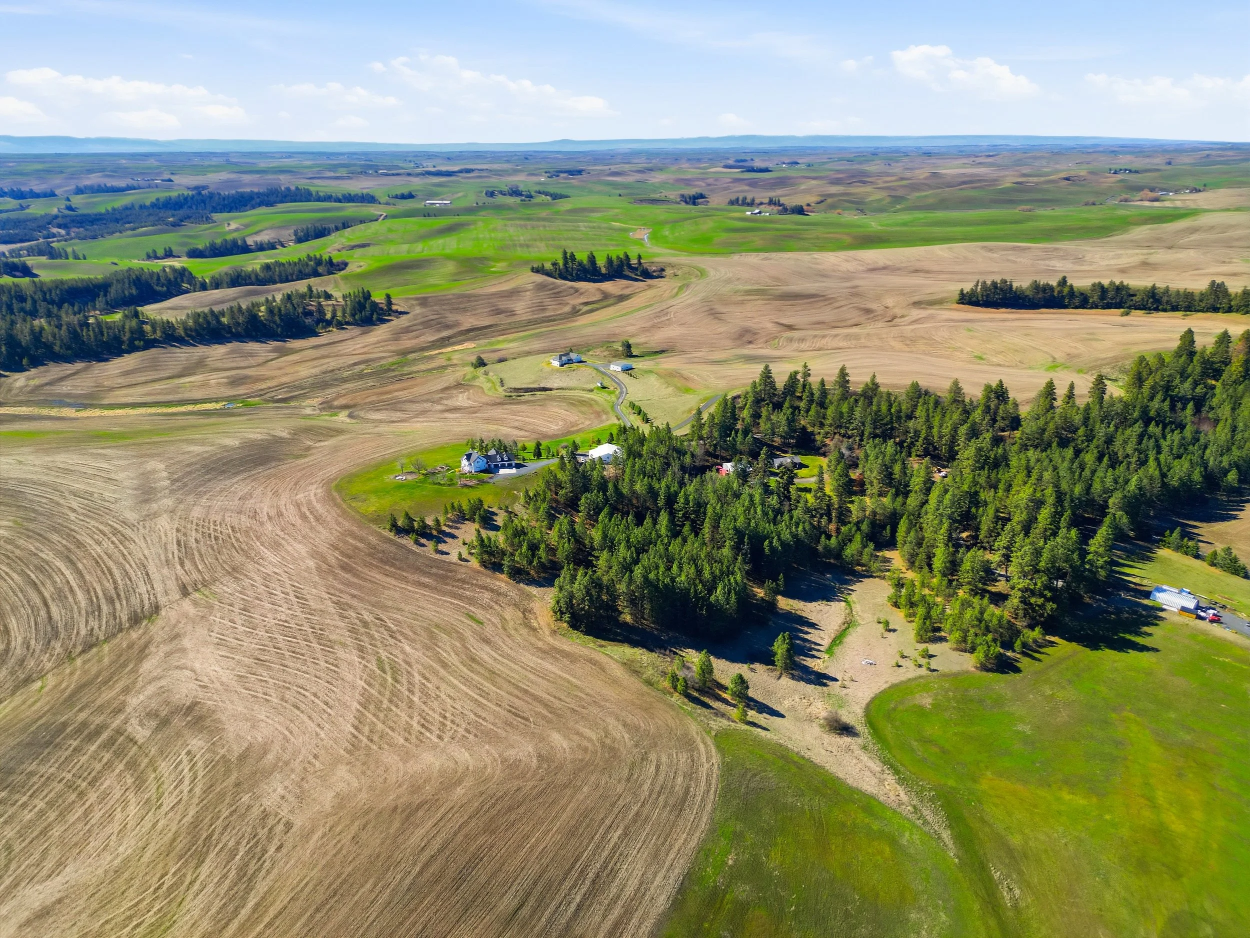 Aerial view of a rural landscape with rolling hills, farmland, scattered houses, and dense forests under a partly cloudy sky.