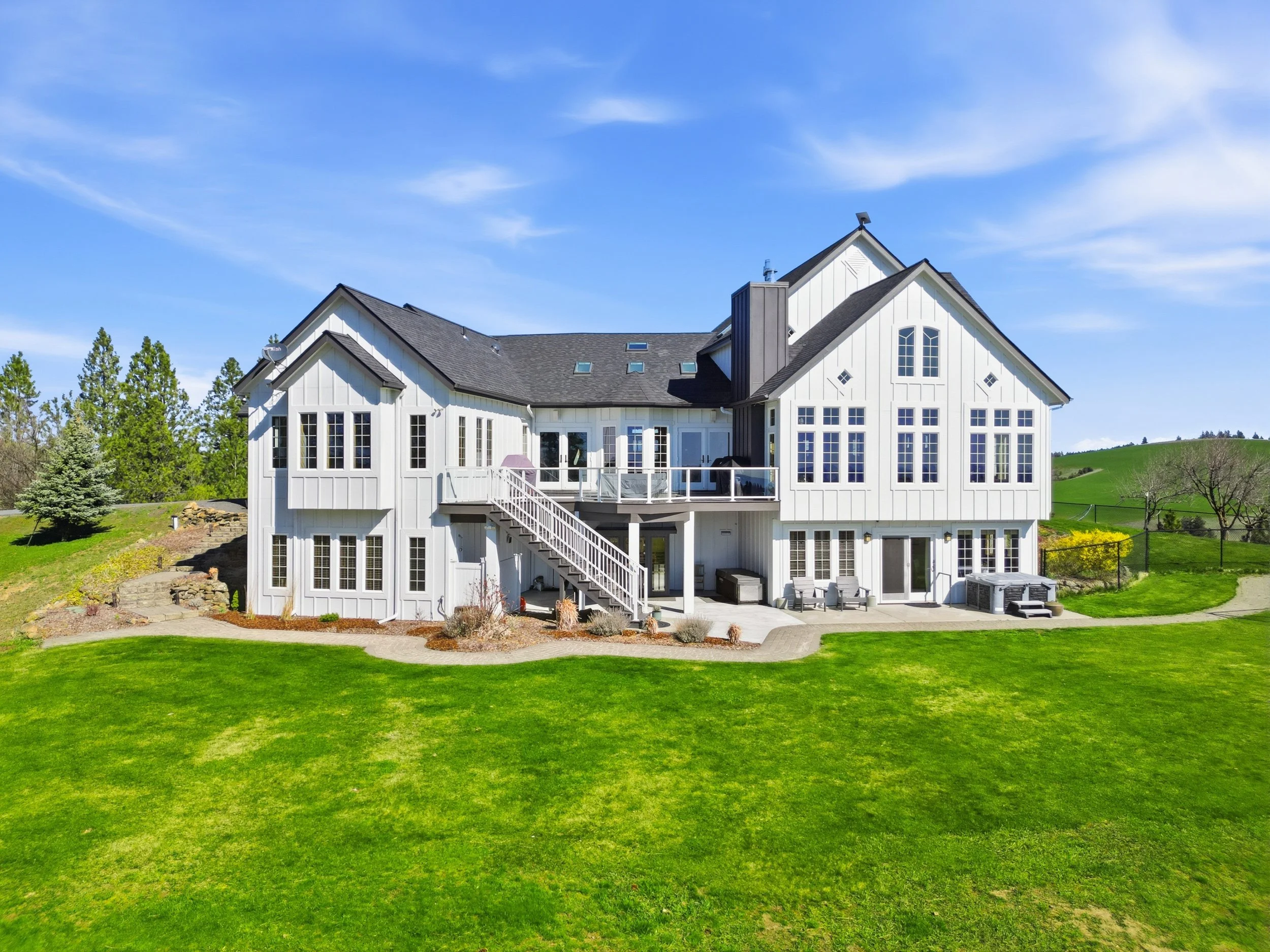 A large white house with multiple windows, a deck, and a sloped roof, surrounded by a well-maintained green lawn and some trees under a blue sky with scattered clouds.
