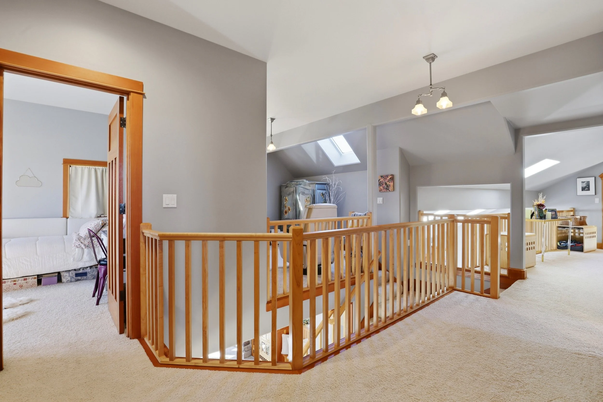 An upstairs area of a house with a beige carpet, wooden railing, and white walls. There are multiple ceiling light fixtures and a skylight. Visible are a small ladder, a sitting area with a vintage wardrobe, and a table with framed photos, a vase, an