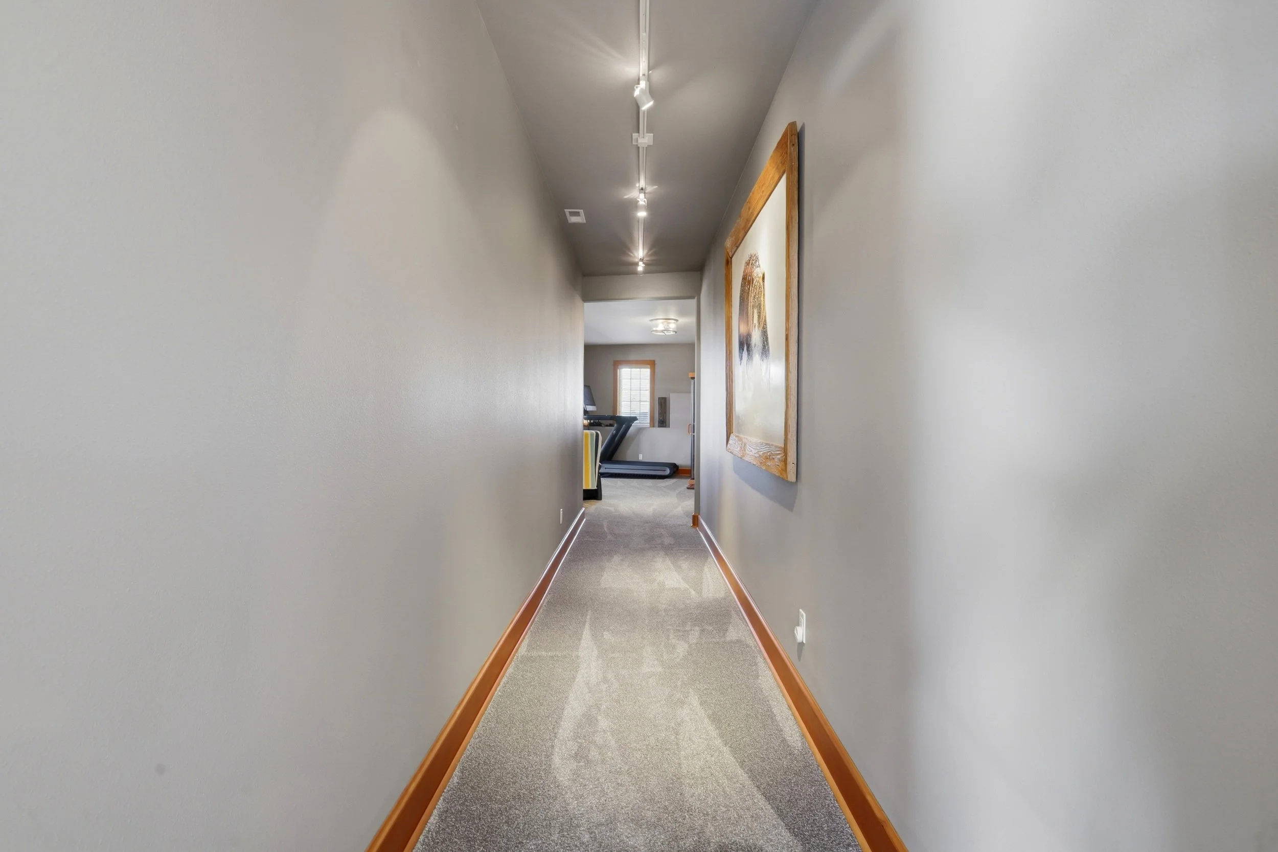 A hallway with gray walls and a wooden baseboard, leading to a living area with a treadmill and a window.