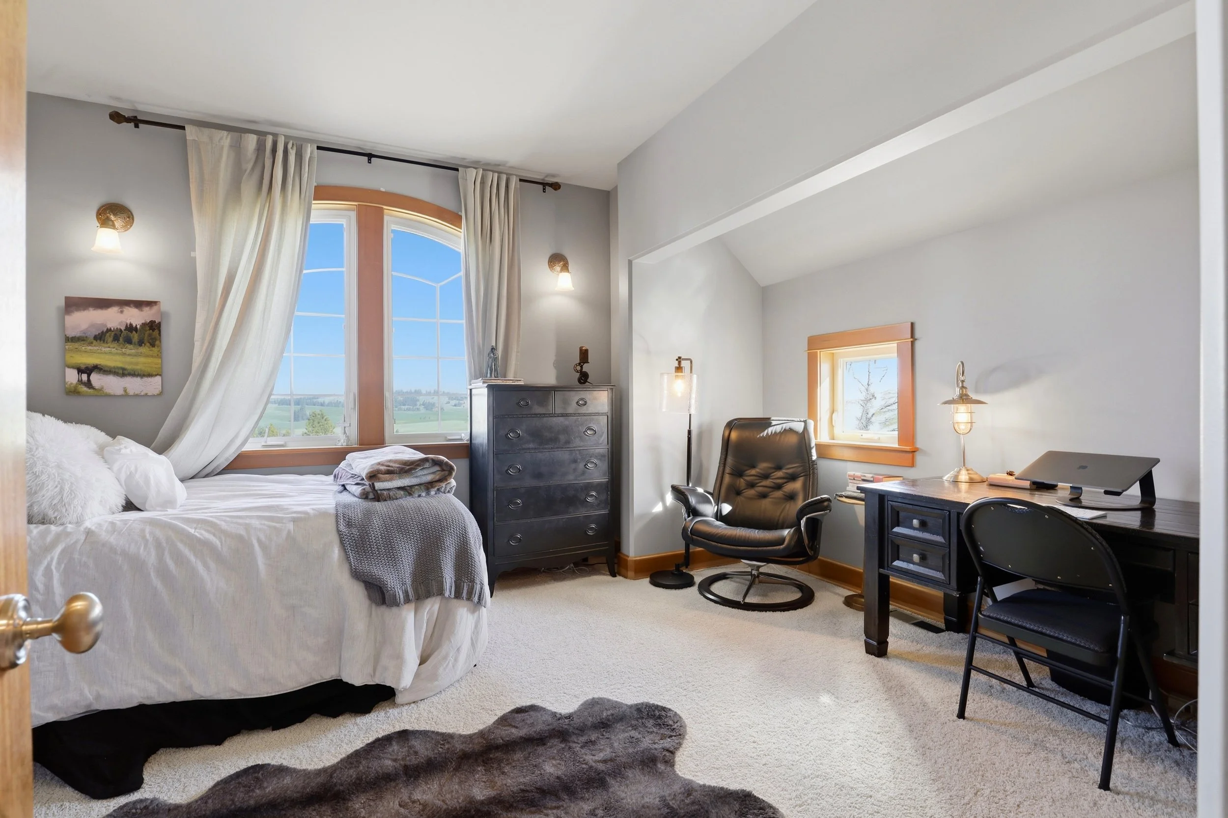 A bedroom with a large window showing a scenic view, a bed with white linen and pillows, a black dresser, a black office chair, a desk with a lamp, a small window, and a dark shaggy rug.