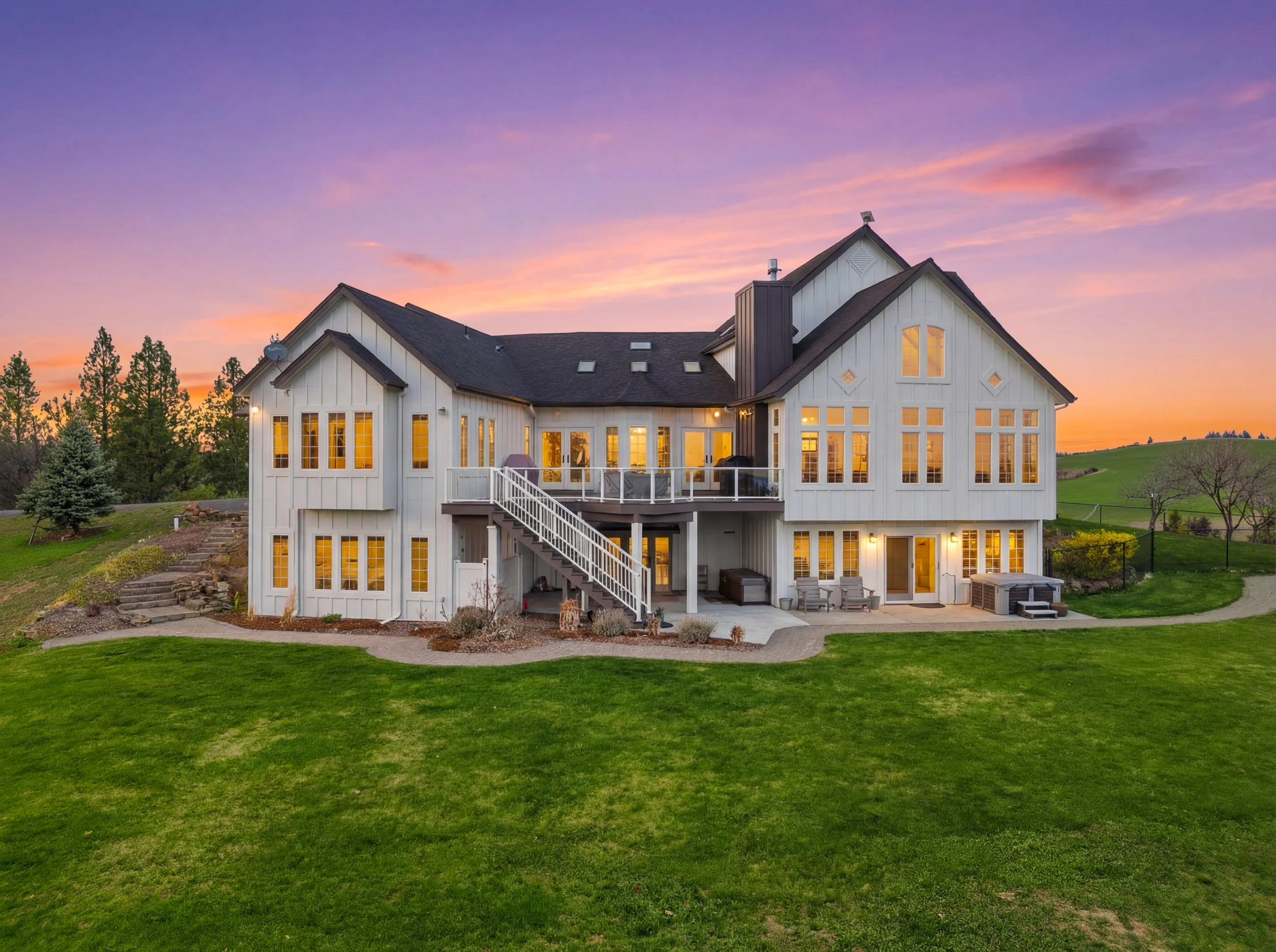 A large white house with multiple gables and large windows glowing with warm light at sunset. The house has a deck with outdoor furniture and stairs leading down to a well-maintained green lawn. In the background, there are trees and rolling hills un