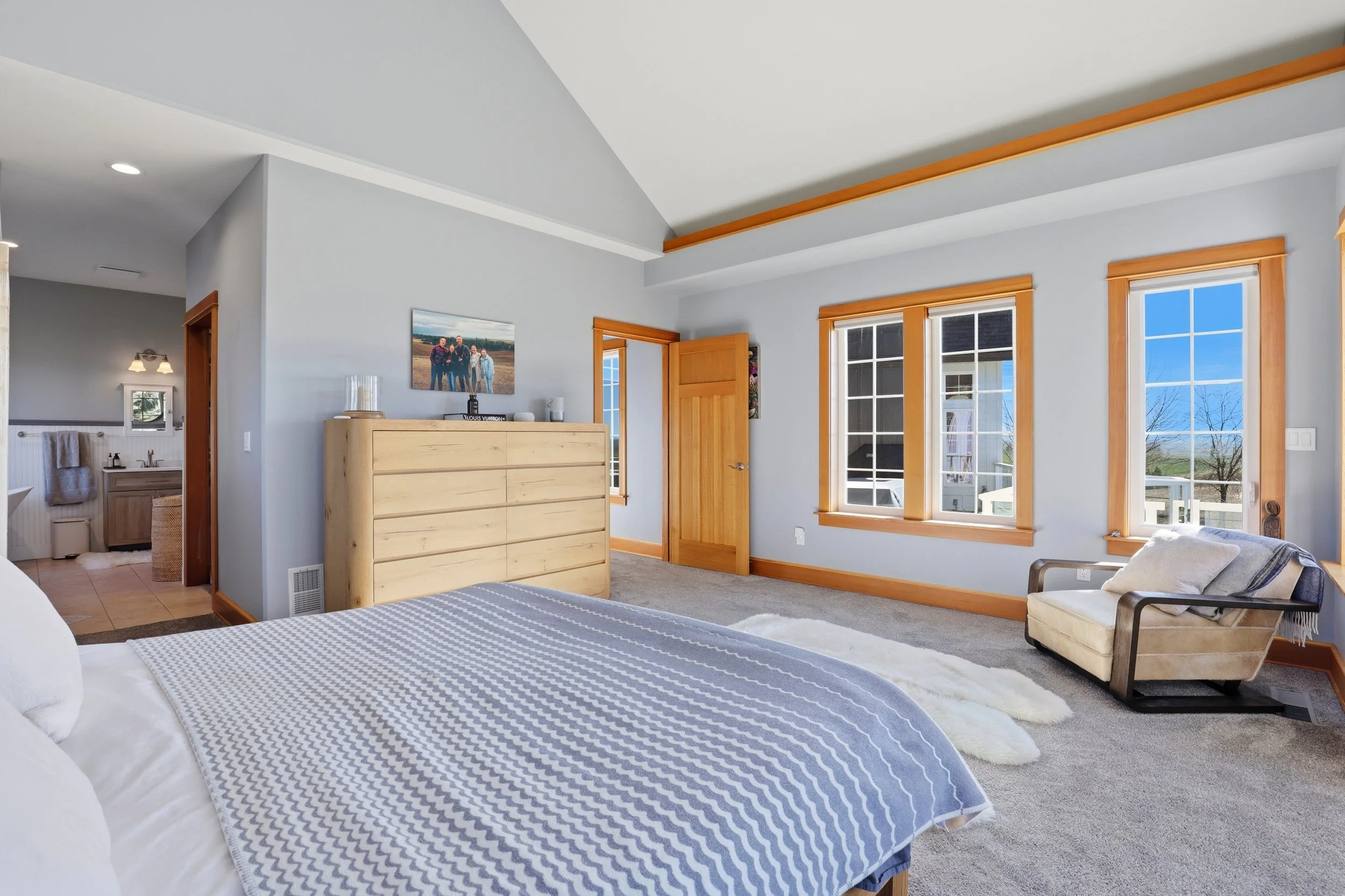 A bright bedroom with light blue walls, wooden window frames, and a view of a clear blue sky outside. There is a bed with a gray-and-white patterned blanket, a beige armchair with a throw blanket, a window with blinds, and a dresser with framed famil