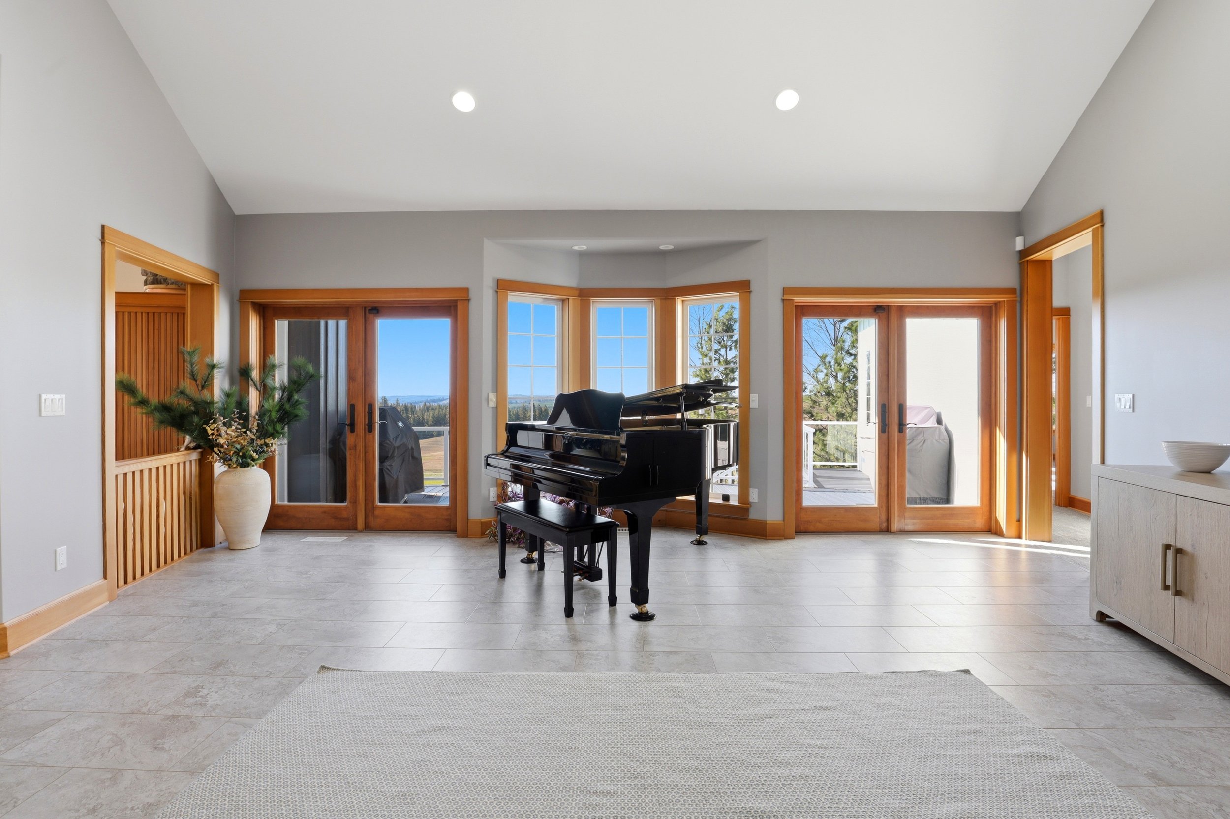 A spacious living room with large windows and glass doors, a black grand piano in the center, a large potted plant to the left, and outdoor view of trees and sky visible through the windows.
