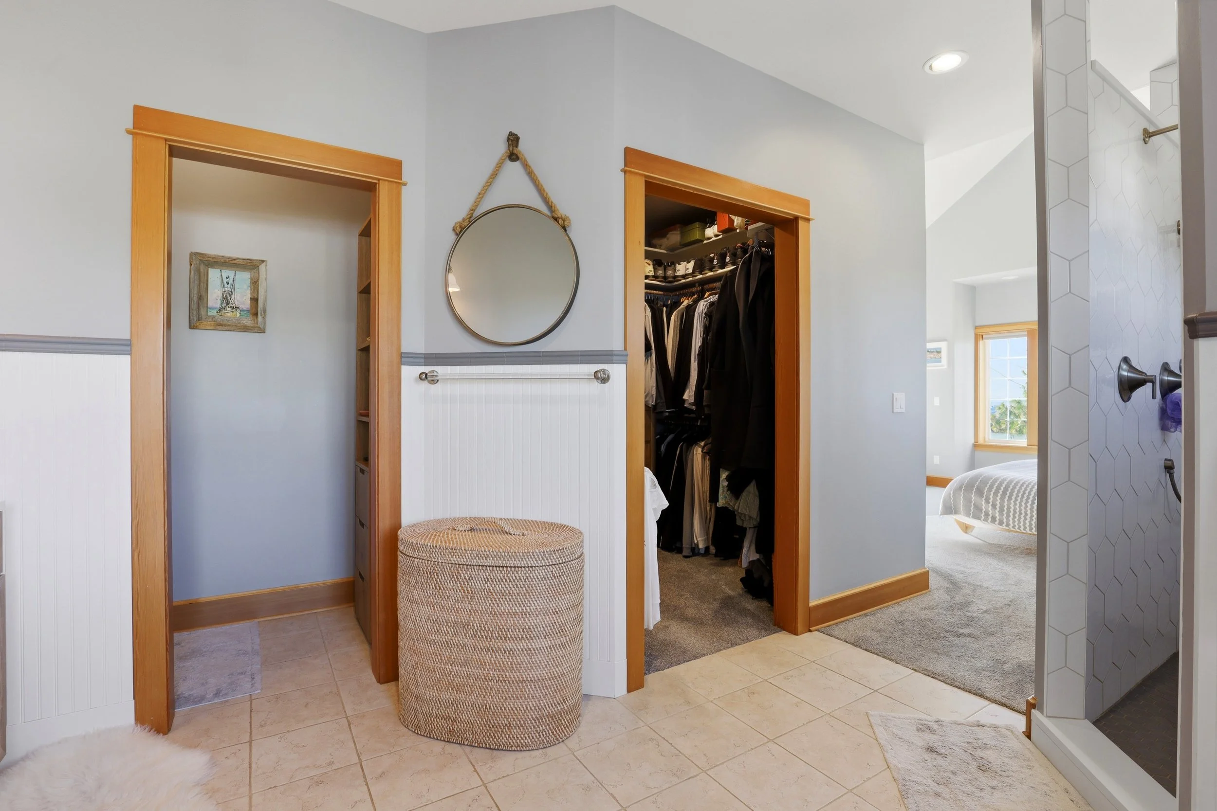 A view of a bedroom with two open closet doors showing hanging clothes, a wall mirror with a rope hanger, and a small ottoman seat in front. The room has beige tiled floors, white beadboard walls with wood trim, and a small painting hanging above one