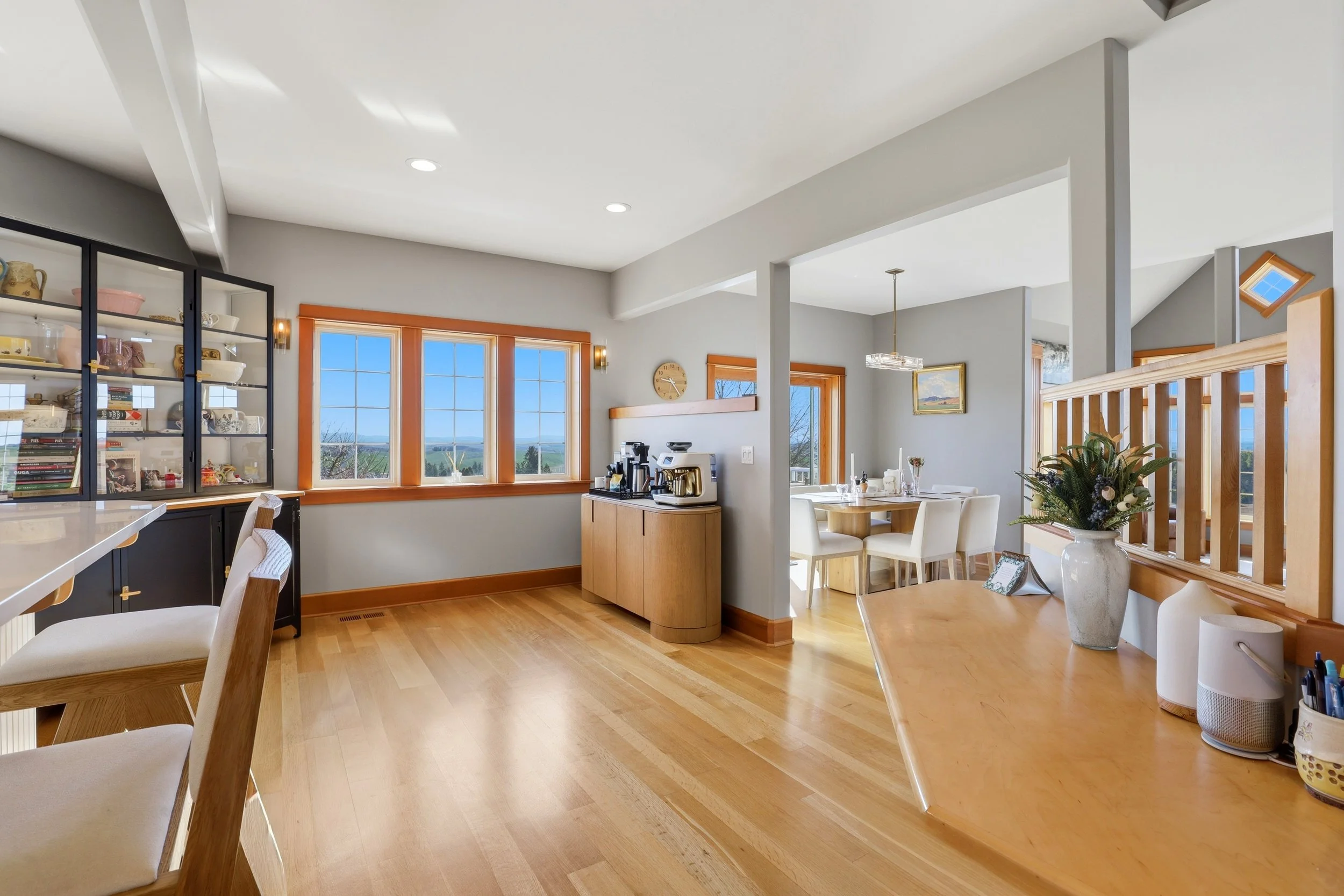 Bright, open kitchen and dining area with large windows, wooden floors, and modern decor, including a coffee station, framed pictures, and a floral arrangement.