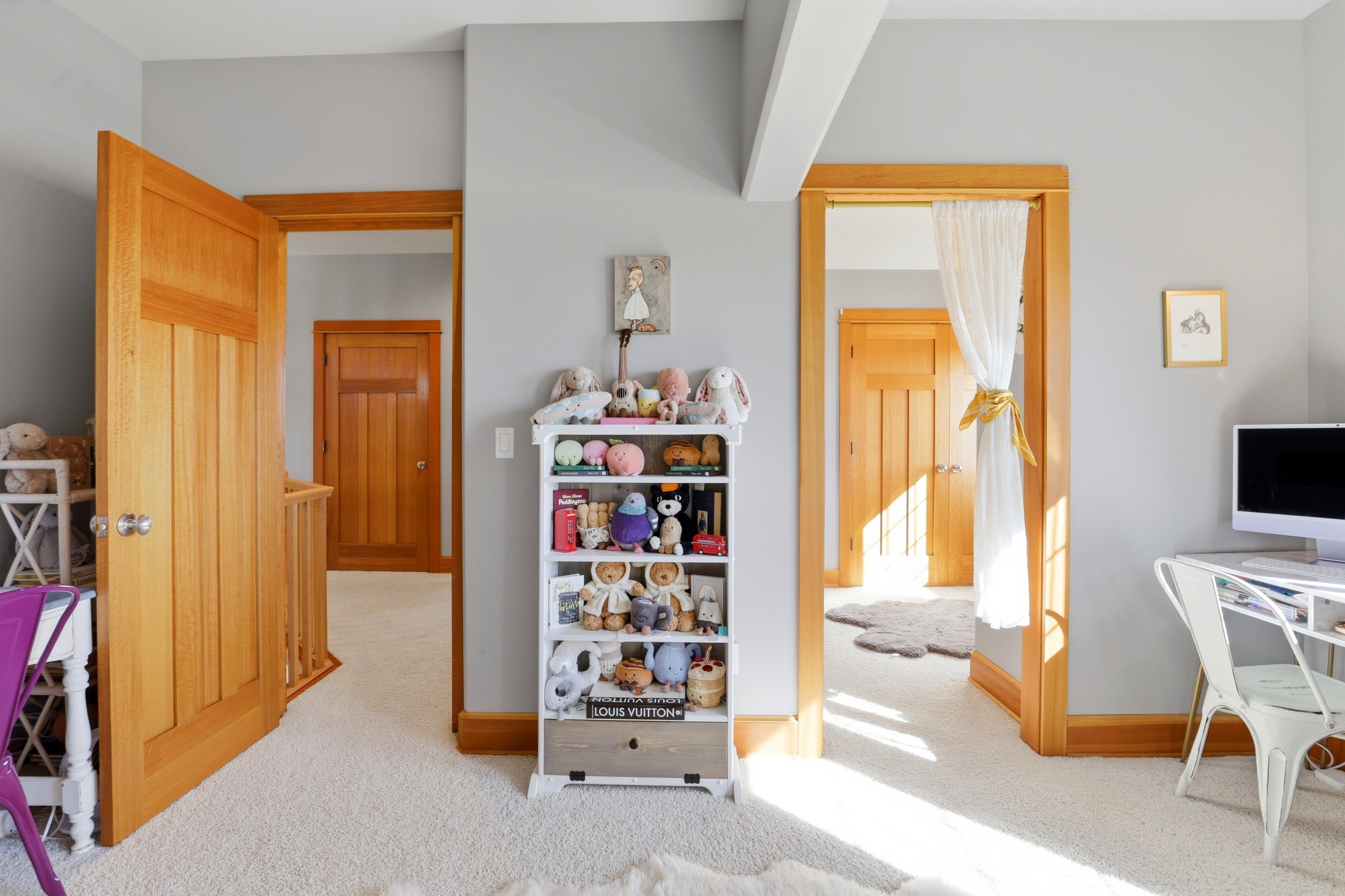 A children's bedroom with a white shelving unit filled with stuffed animals and toys, wooden door frames, and a window with a white curtain tied back, letting sunlight in.