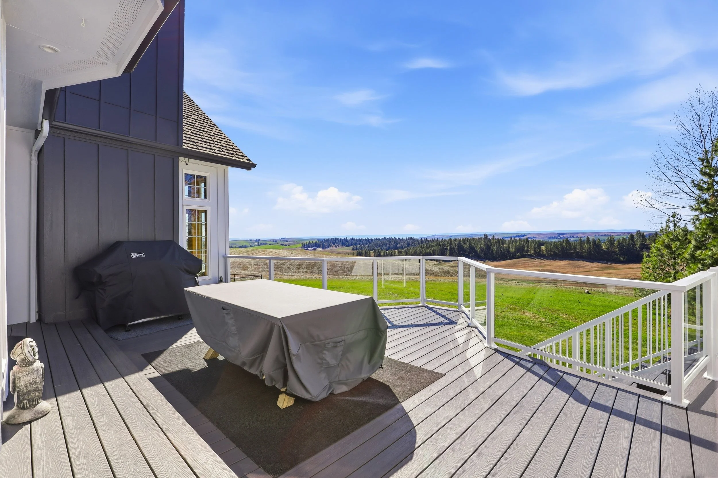 A spacious balcony with wooden flooring and white railing overlooking a scenic rural landscape with fields, trees, and a clear blue sky.