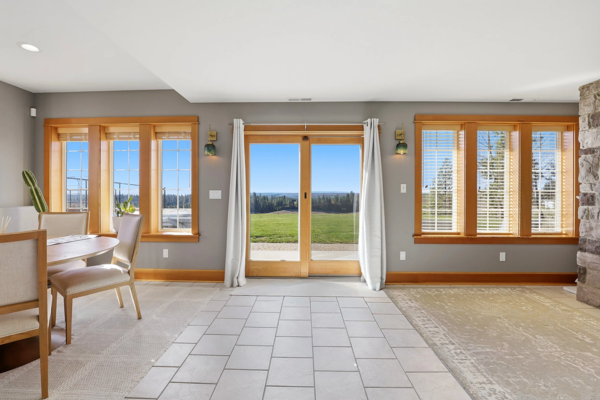 A living room with large windows and a sliding glass door showing a view of green grass and trees outside. There are curtains, a dining table with chairs, a potted plant, and a mix of tile and rug flooring.
