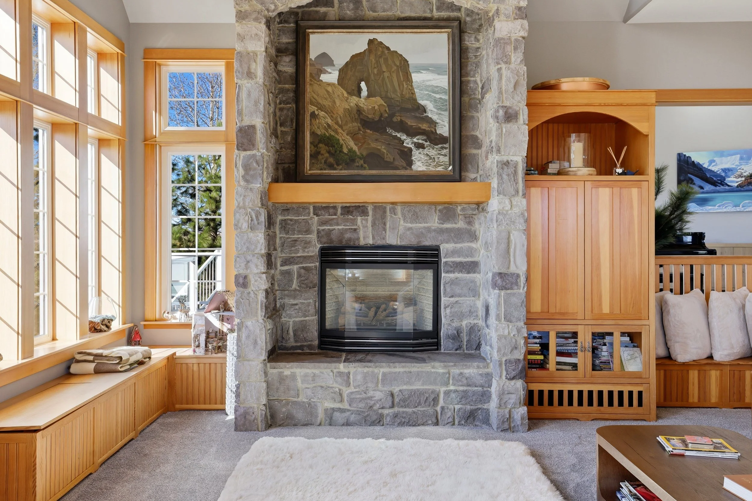Living room with large window, stone fireplace with artwork of a rocky coastal scene, wooden built-in shelves, and cozy cushions on a beige sofa.