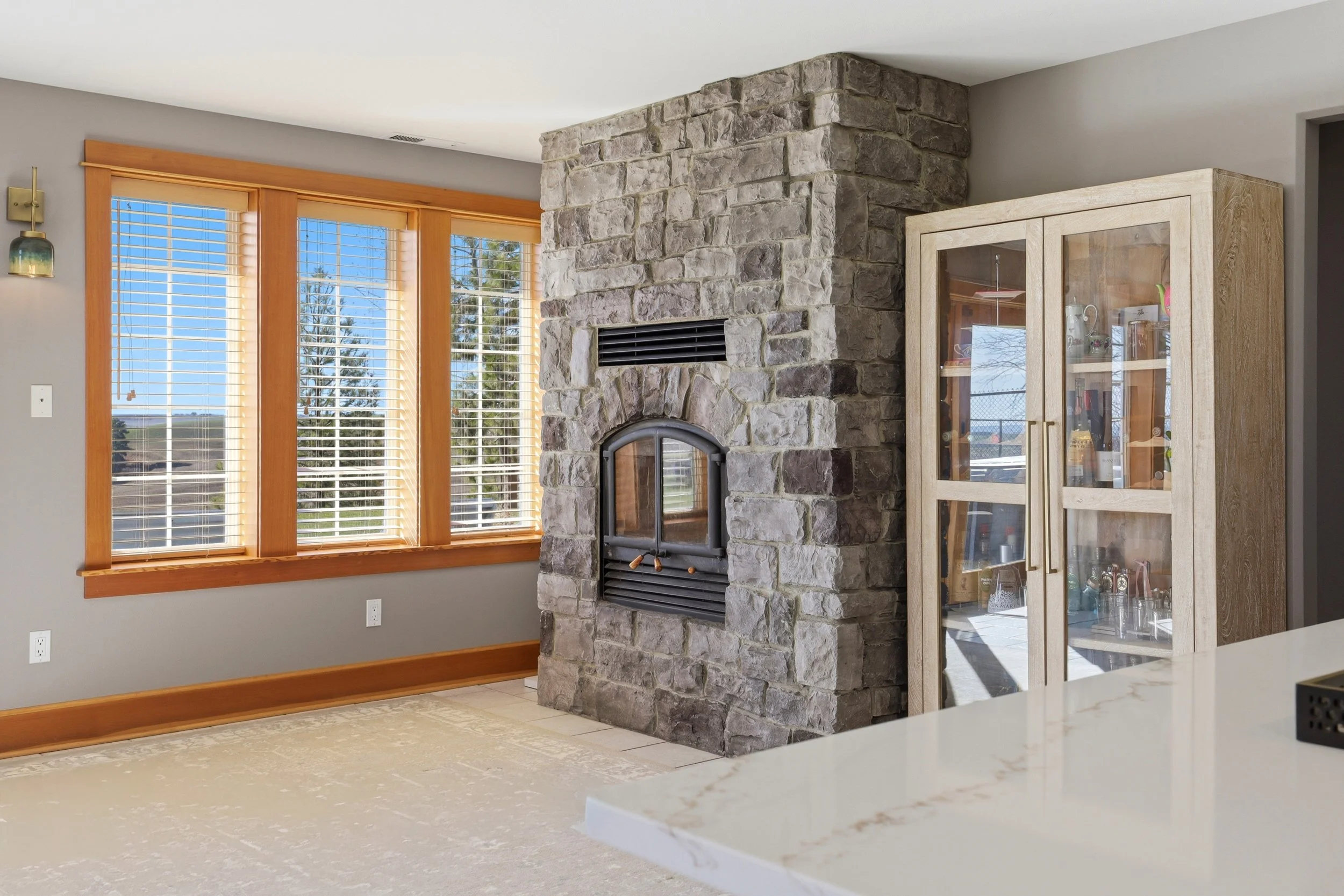 Living room with large window with wooden blinds, stone fireplace, and wooden cabinet with glass doors containing decorative items.