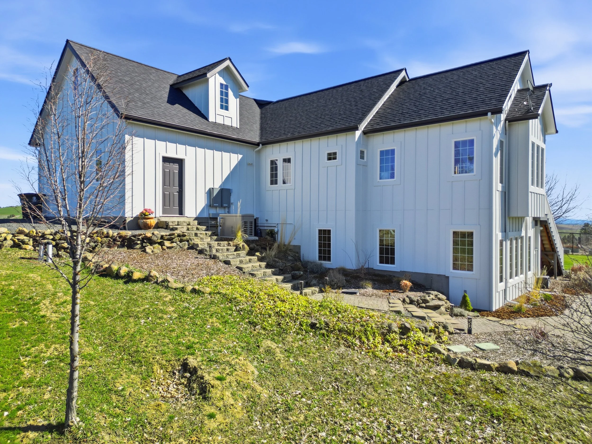 A white house with black roofing shingles, multiple windows, a gray front door, and a small porch with stairs leading up to it. There is a grassy yard with a small tree and a rock garden in front, under a clear blue sky.