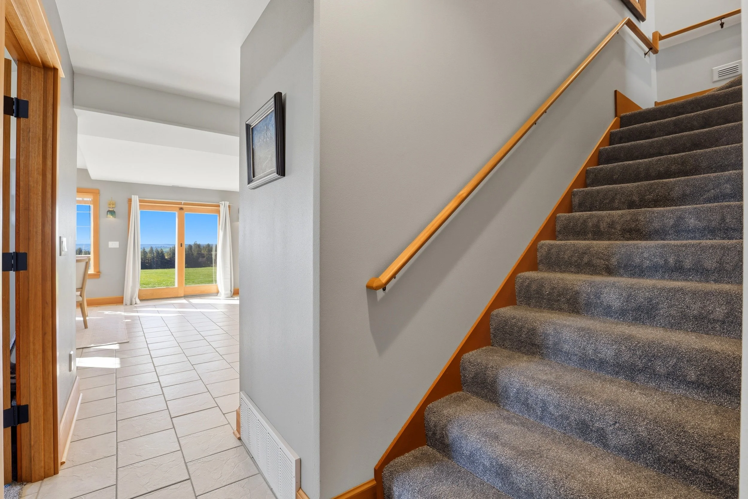 View of a staircase with gray carpet, wooden handrail, and beige walls, adjacent to a bright living area with large windows and sliding glass doors showing a green outdoor landscape.