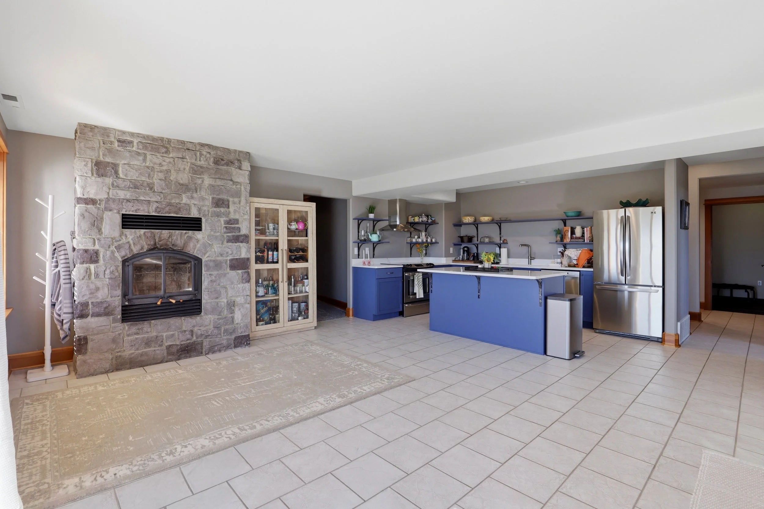 A kitchen with a stone fireplace, blue cabinets, stainless steel refrigerator, open wall shelves, and a beige area rug on tiled floor.
