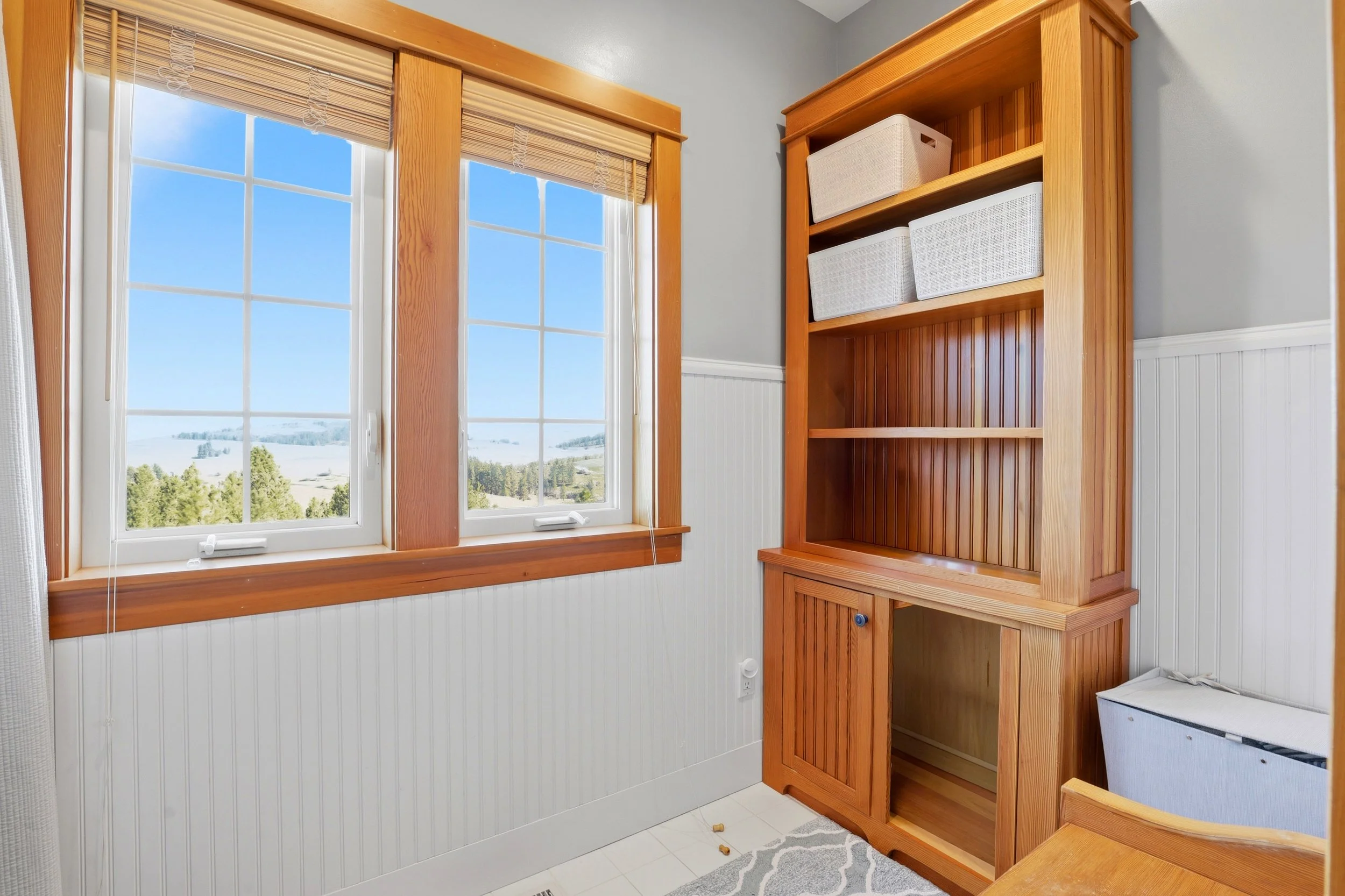 A room with two windows with blinds, a tall wooden shelving unit with baskets, and a small white cabinet.
