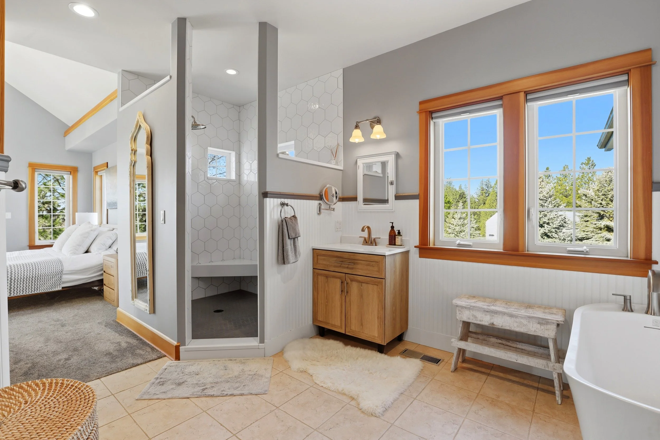 Interior view of a modern home bathroom with a large window, wooden trim, a vanity with a sink, a mirror, and a shower with white hexagonal tiles, with a glimpse of a bedroom in the background.