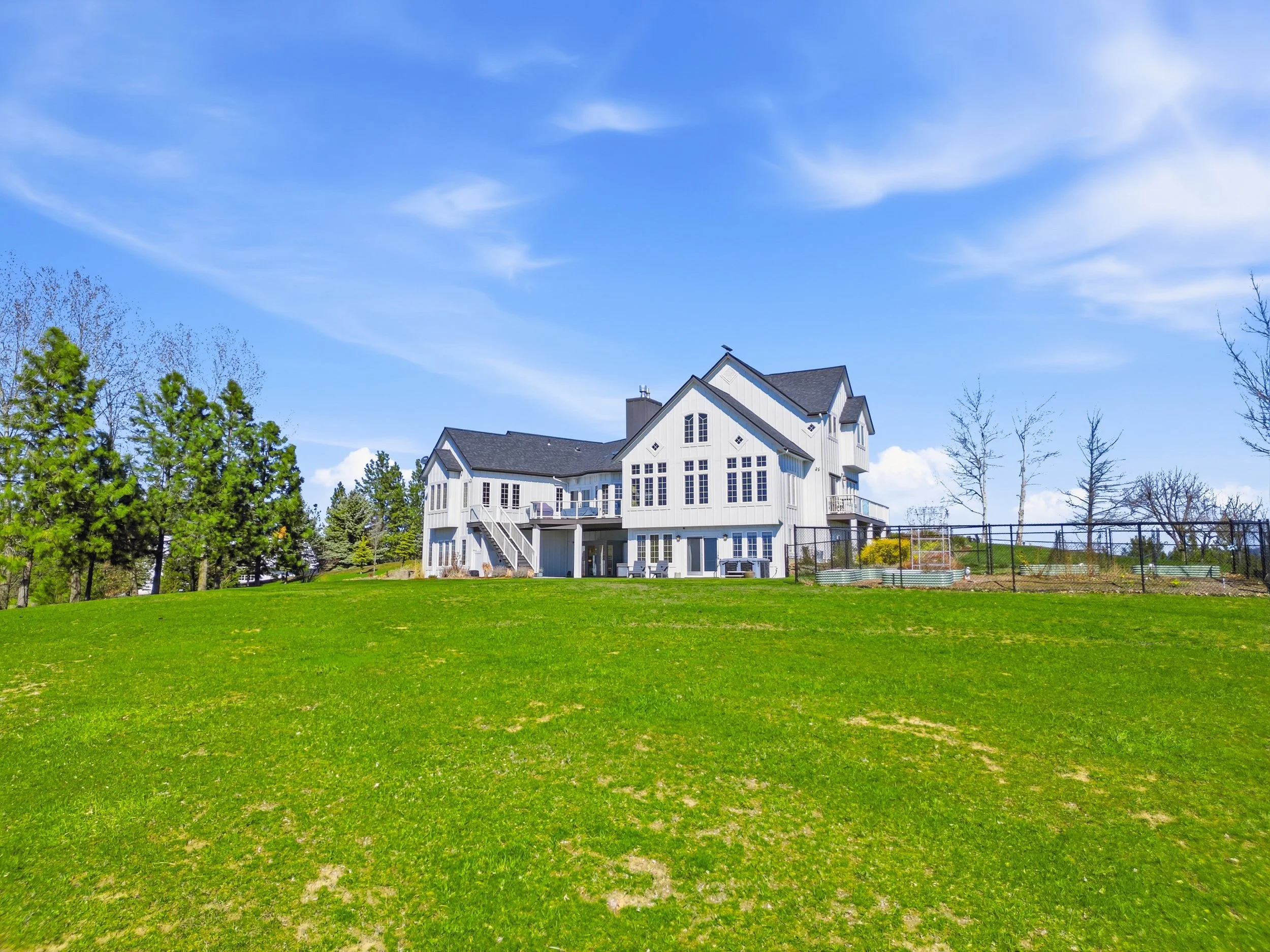 Large white house with multiple gabled roofs and large windows, situated on a lush green lawn with several trees and a black fence in the background, under a bright blue sky with scattered clouds.