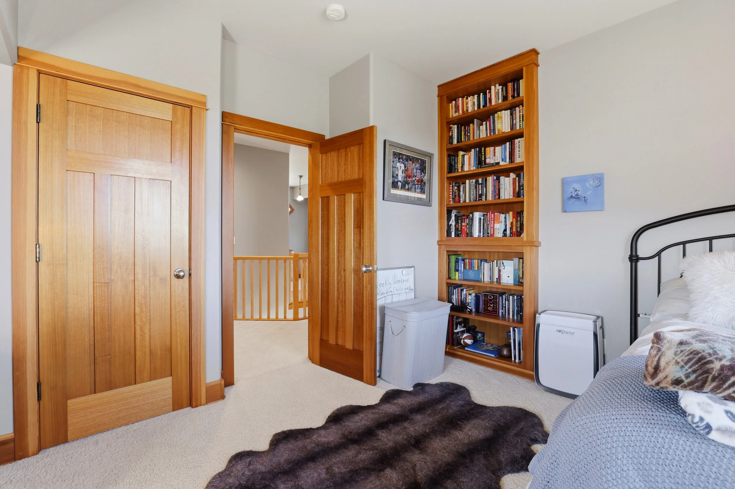 A bedroom with two wooden doors, a tall wooden bookshelf filled with books, a framed picture on the wall, a striped armchair with a pillow, a furry brown rug on beige carpet, and a white air purifier.