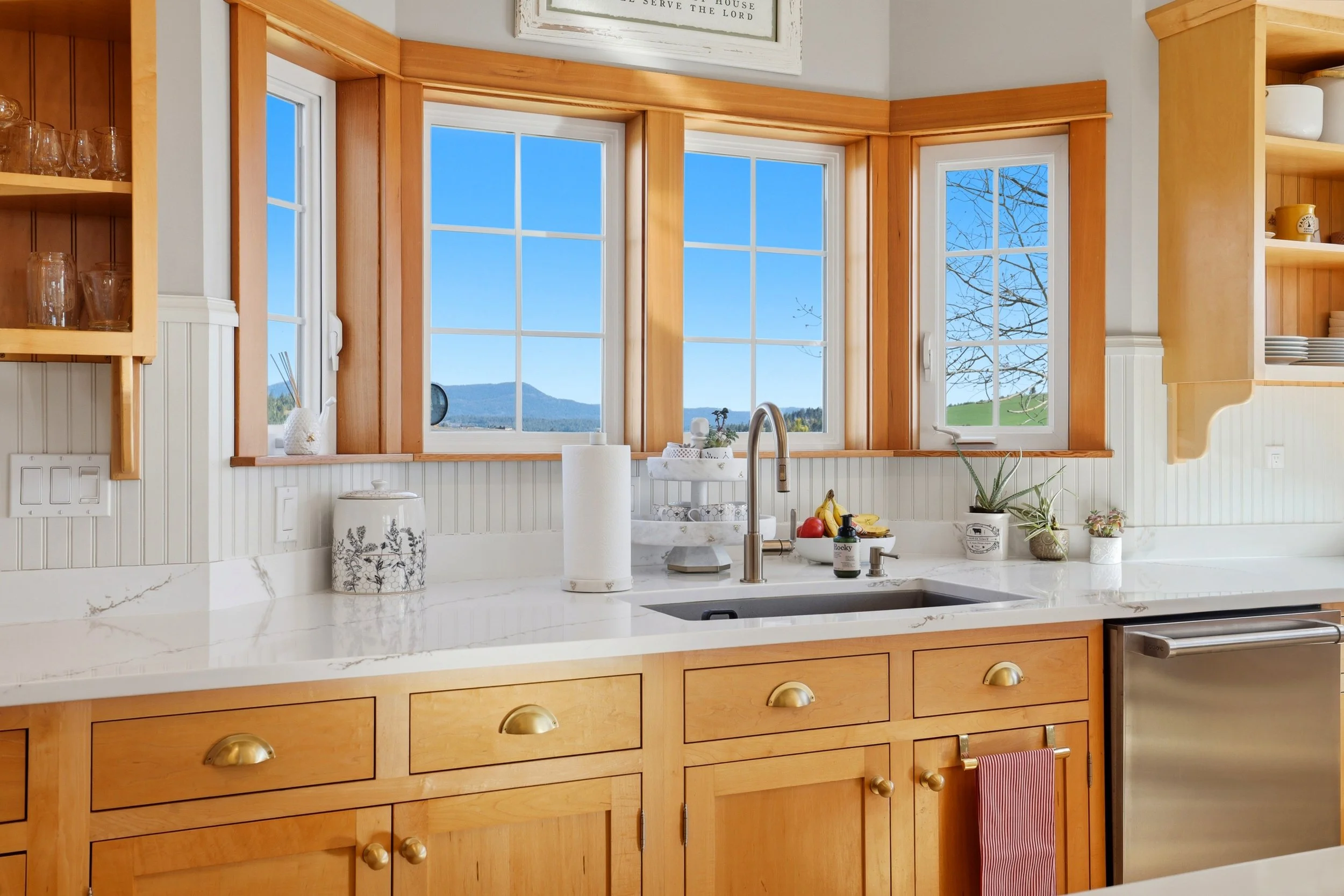 A kitchen with large windows showing a scenic view of mountains and trees outside. The kitchen has wooden cabinets, a white marble countertop, and various kitchen items, including a paper towel roll, a vase, fruit, and plants.