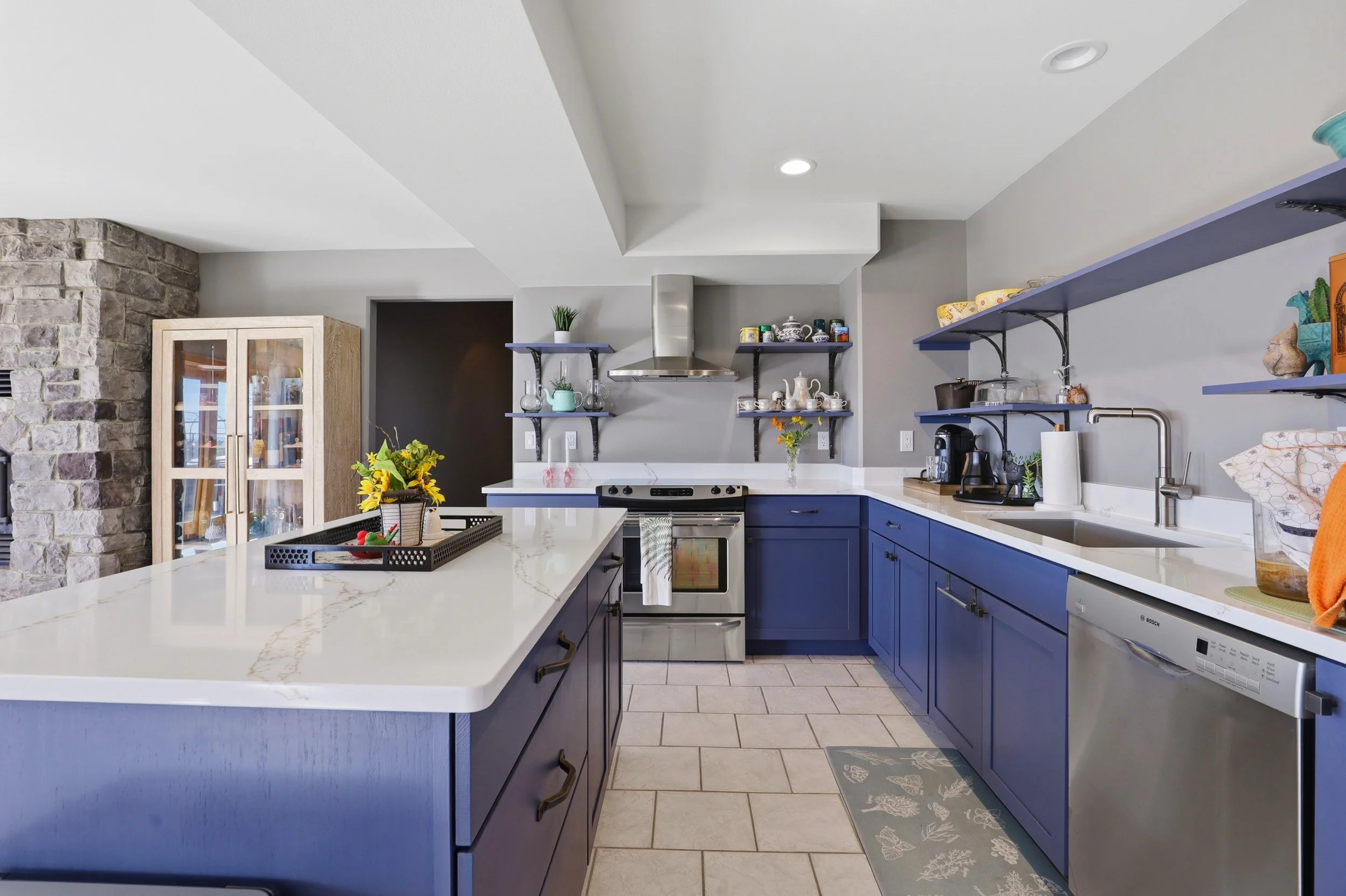 Modern kitchen with blue cabinets, white countertops, open shelves with dishes, a stove with a stainless steel range hood, a glass-front cabinet, and various decorative items and plants.
