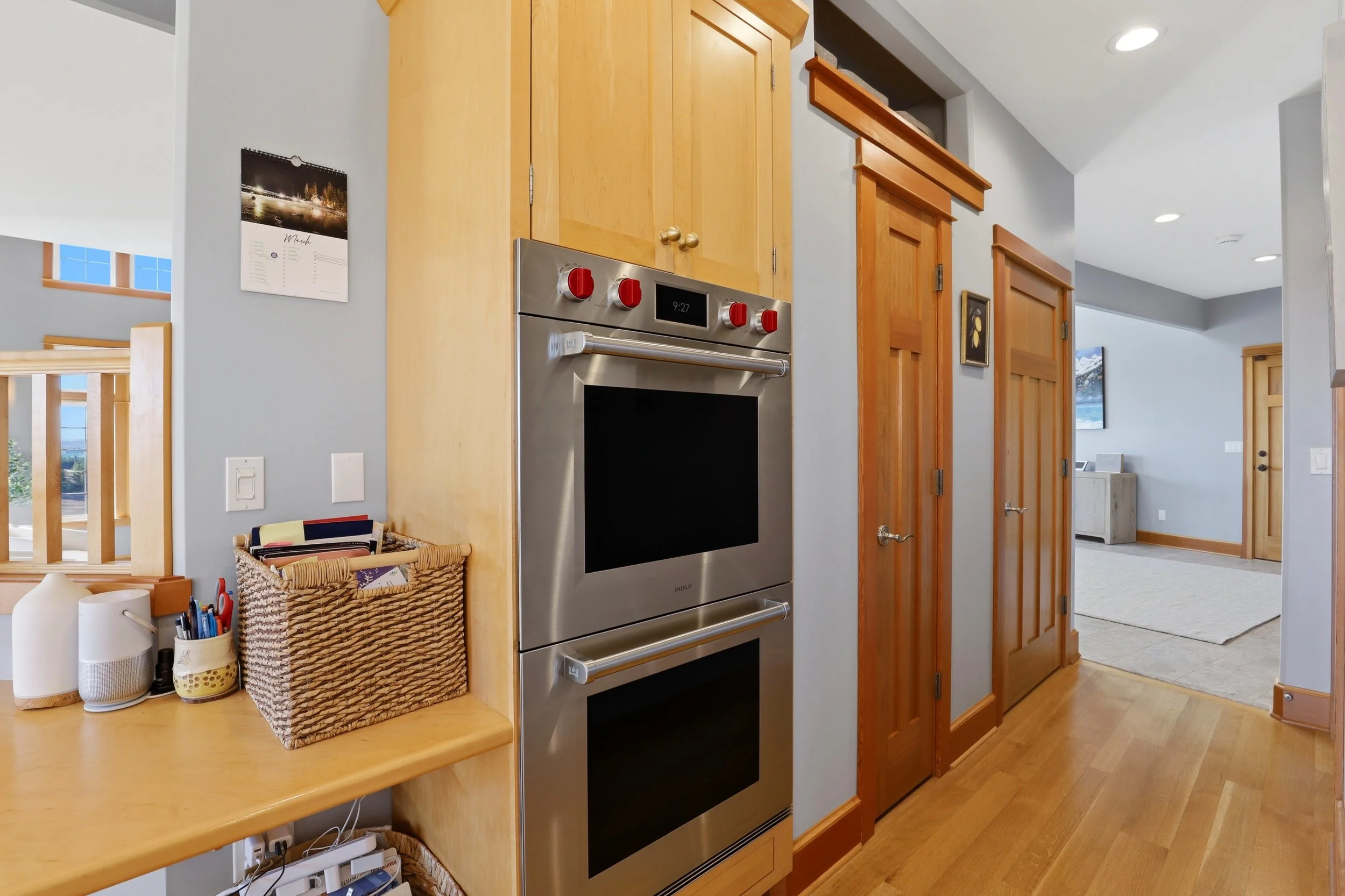 A modern kitchen with a stainless steel oven installed in wooden cabinetry. To the left, there is a wooden countertop with various items like paper towels, a basket filled with miscellaneous objects, and writing utensils. The space is bright with nat