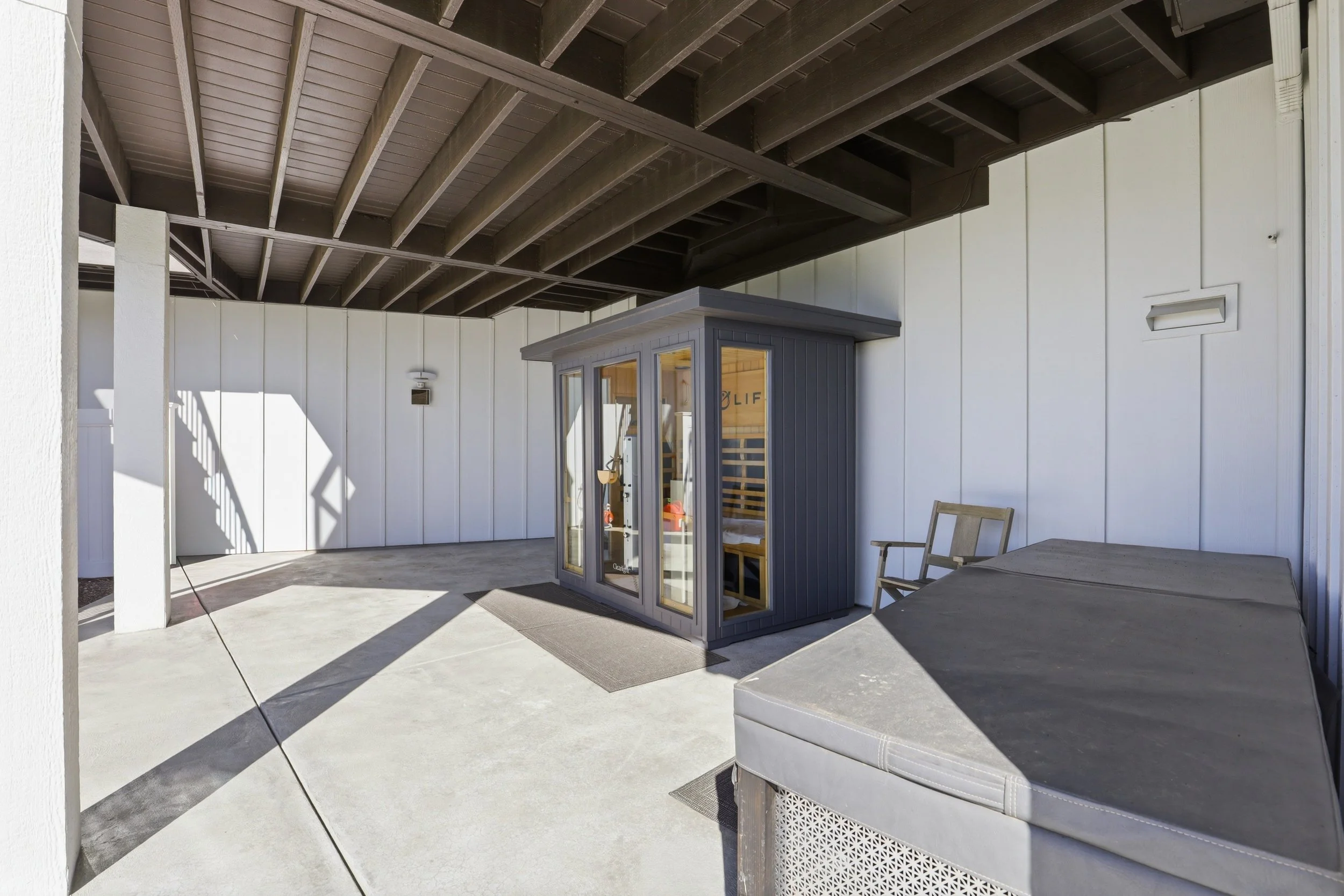 An outdoor patio area under a dark wooden ceiling with white walls, featuring a small gray outdoor storage shed with glass doors, a table, and chairs.