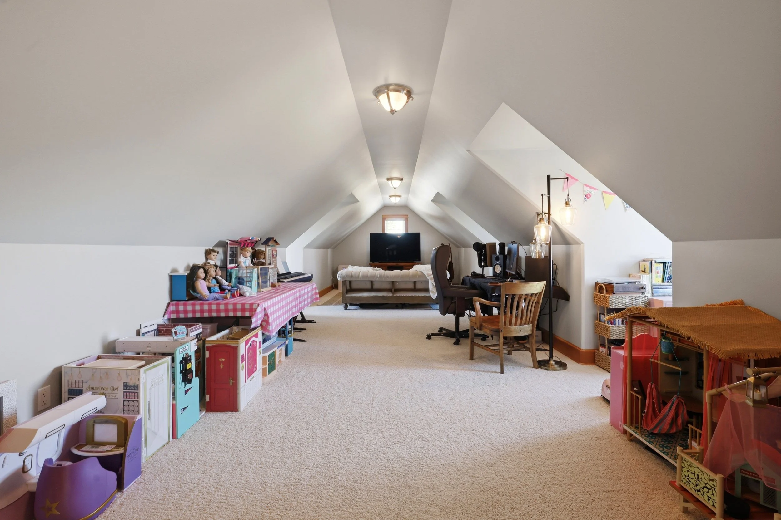 A spacious attic room with sloped ceilings, carpeted floor, and various furniture. On the left, there are dolls and toy furniture aligned on a pink checkered tablecloth. In the center, a black TV is situated in front of a window, accompanied by a bei