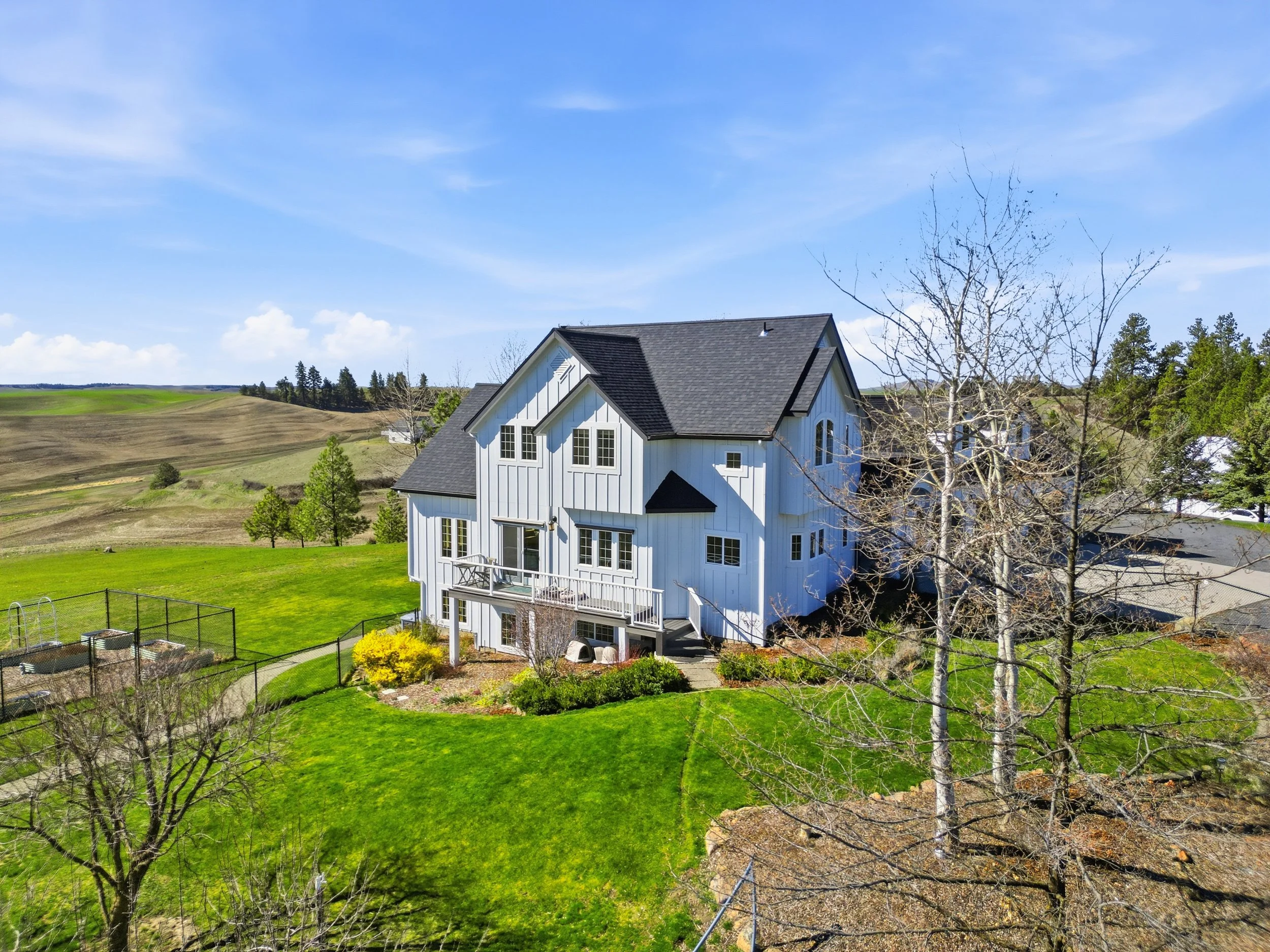 A large white house with blue trim and multiple windows is situated on a lush green lawn, with a fenced-in backyard. The yard features trees, some with blossoming yellow flowers, and a patio area. In the background, rolling farmland and a line of tre