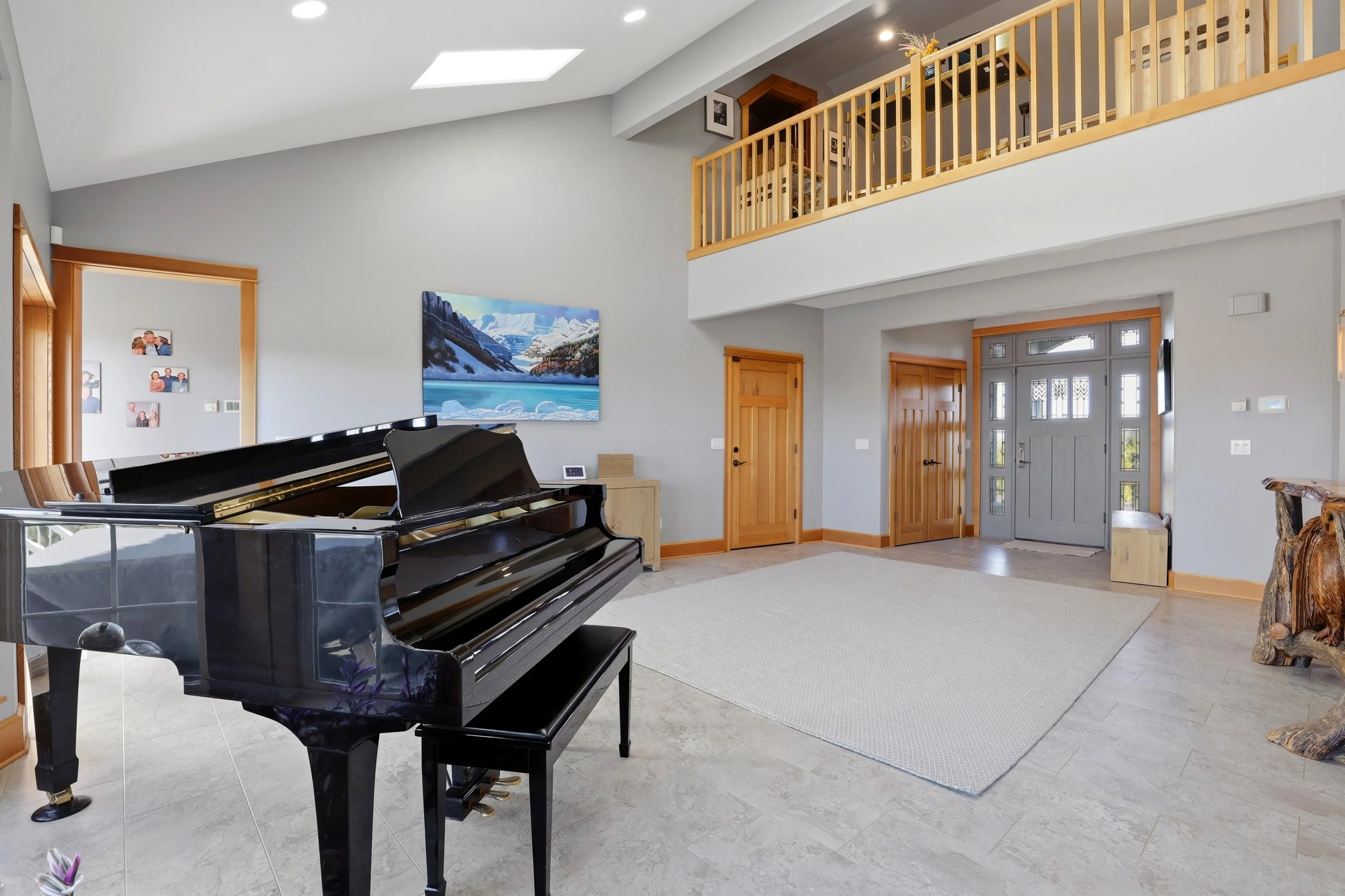 Living room with a black grand piano, a beige area rug, wooden door frames, a wall art painting of mountains and water, and a two-story ceiling with a skylight.
