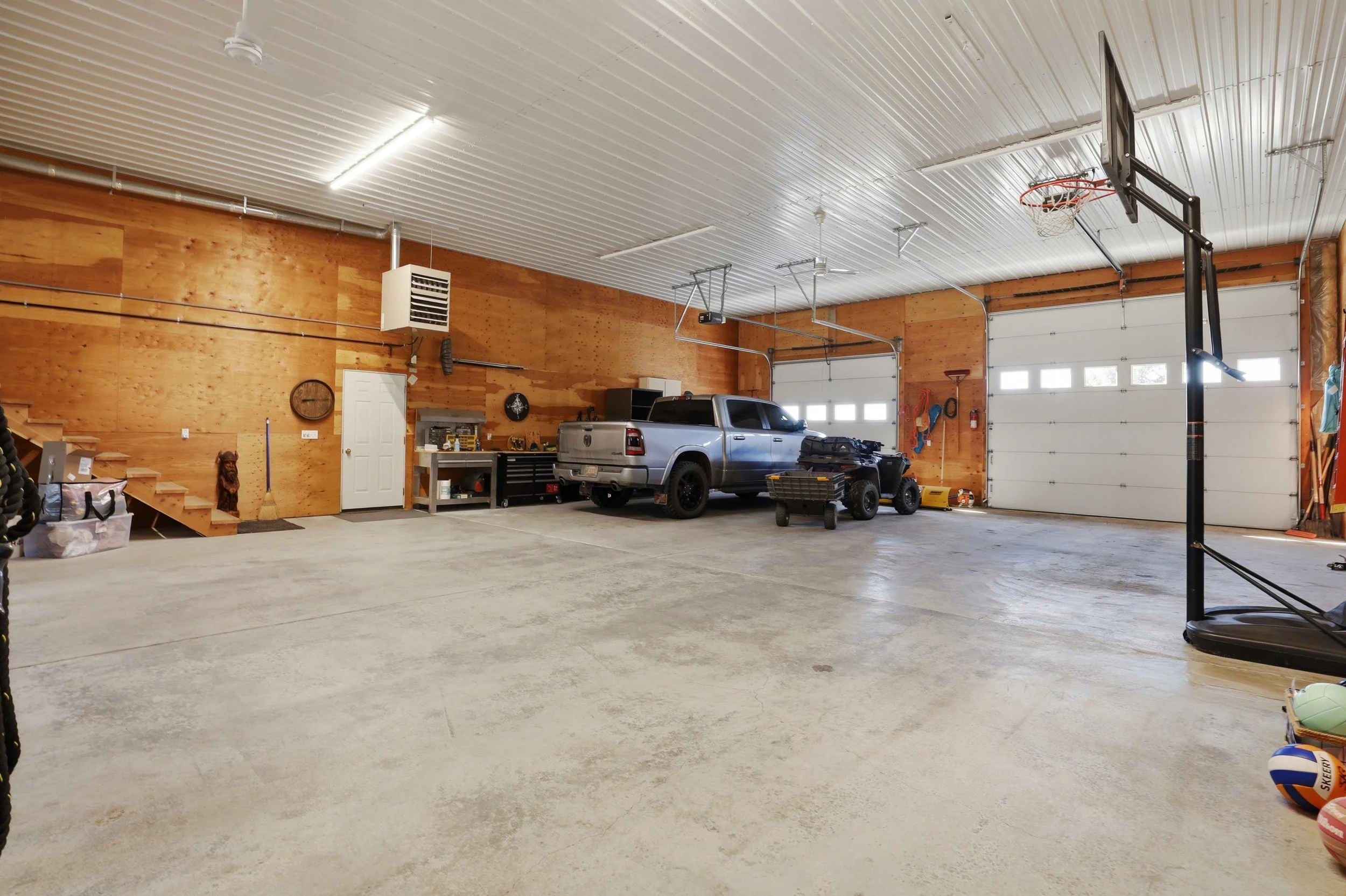 A spacious garage with a concrete floor, wooden paneled walls, and a white ceiling. There is a silver pickup truck and a small ATV parked inside, along with various tools and equipment. A basketball hoop is attached to a black stand, and several larg