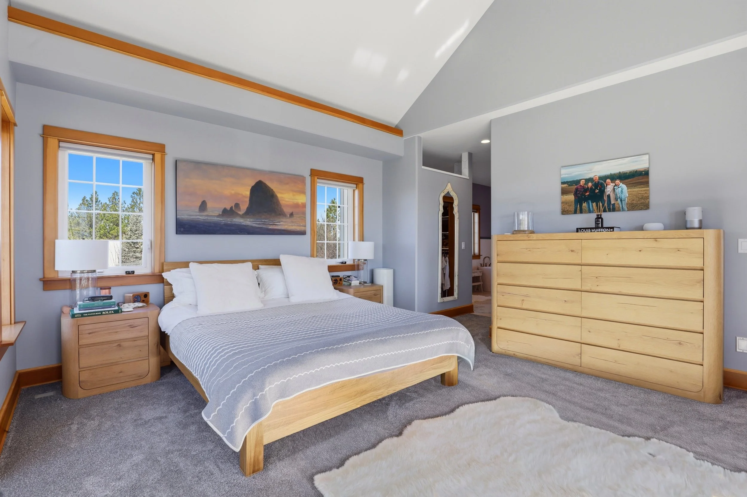 A bedroom with a wooden bed frame and white bedding, two nightstands with table lamps, windows with wooden trim, wall art depicting a sunset and ocean scene, a large dresser, and a fluffy white rug on a gray carpeted floor.