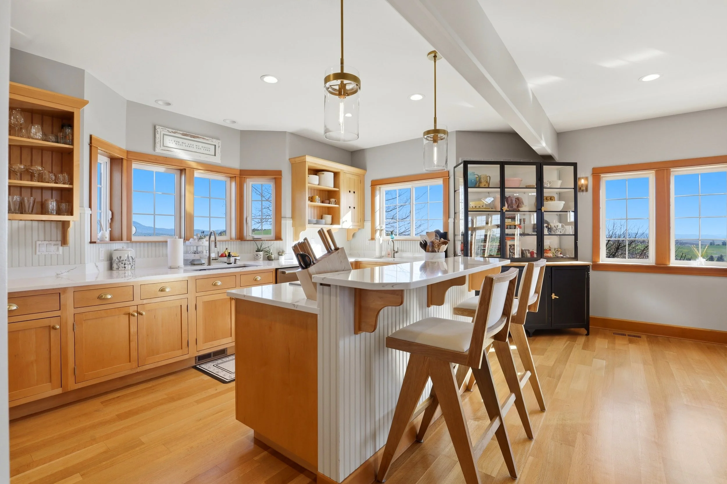 Bright kitchen with wooden cabinets, white countertops, open shelving, black glass display cabinet, kitchen island, bar stools, and large windows showing a scenic outdoor view