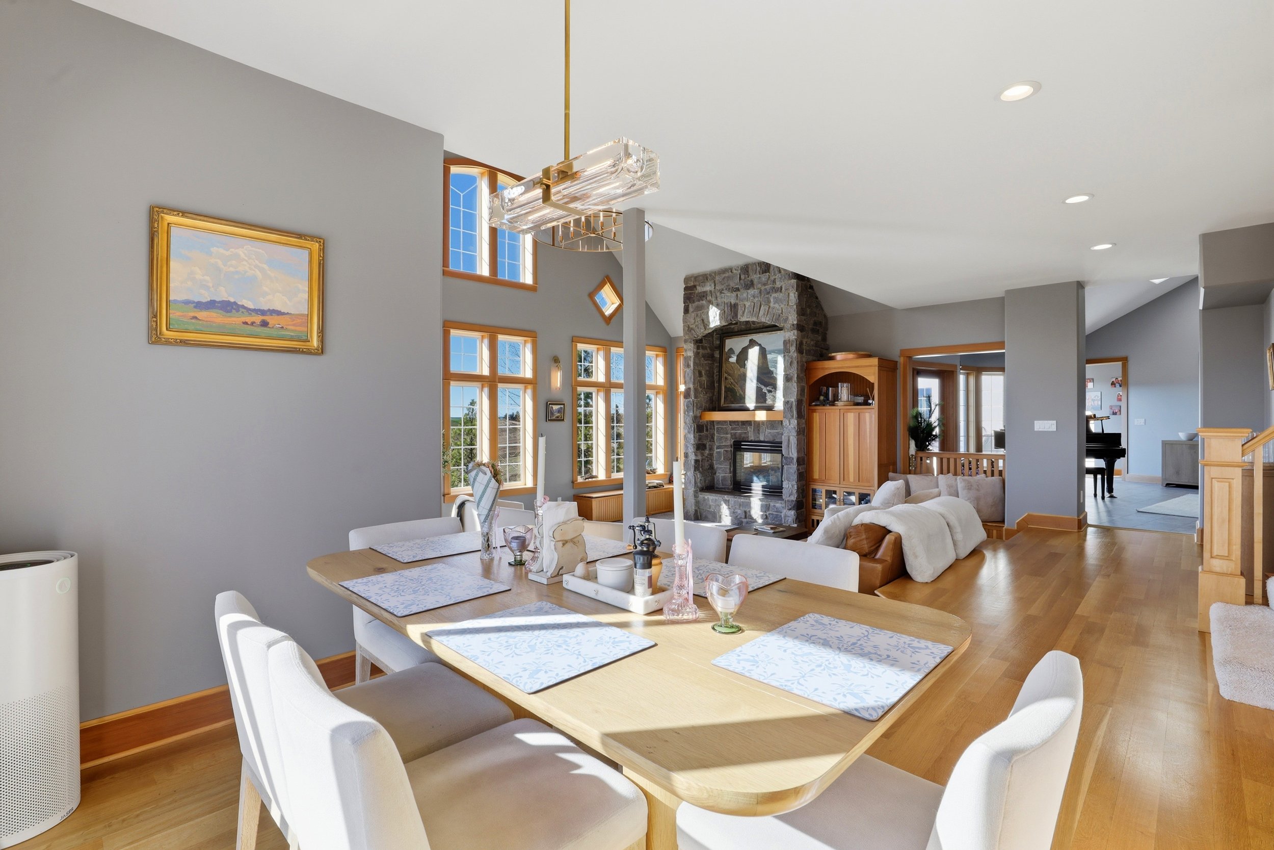 A spacious, modern living and dining area with large windows, a stone fireplace, a wooden dining table with white chairs, and a mix of contemporary and rustic decor.