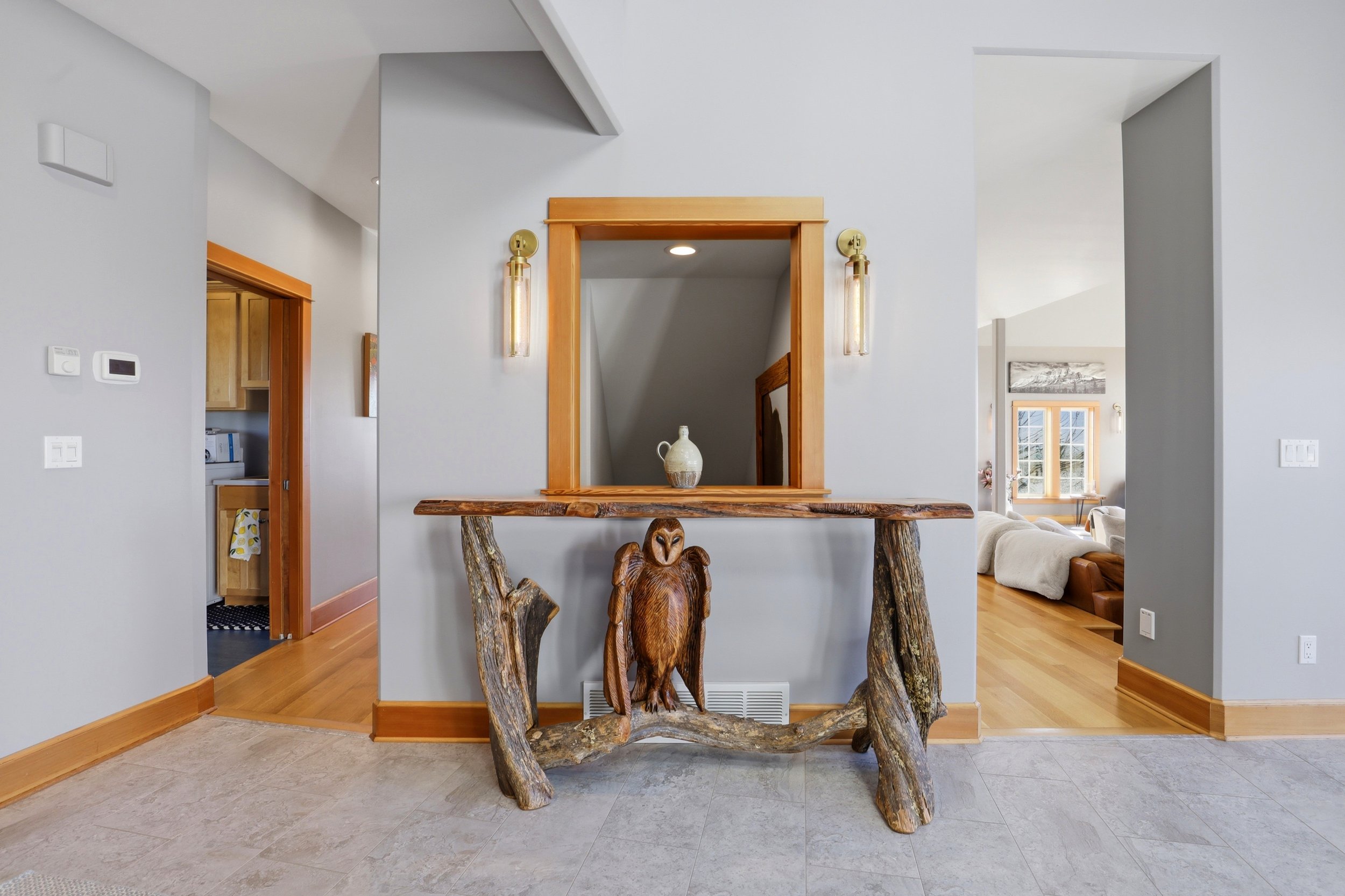 A rustic wooden console table with carved animal figures, including a prominent owl, set against a light gray wall with wooden trim. There are wall-mounted brass sconces and a small window visible in the background.