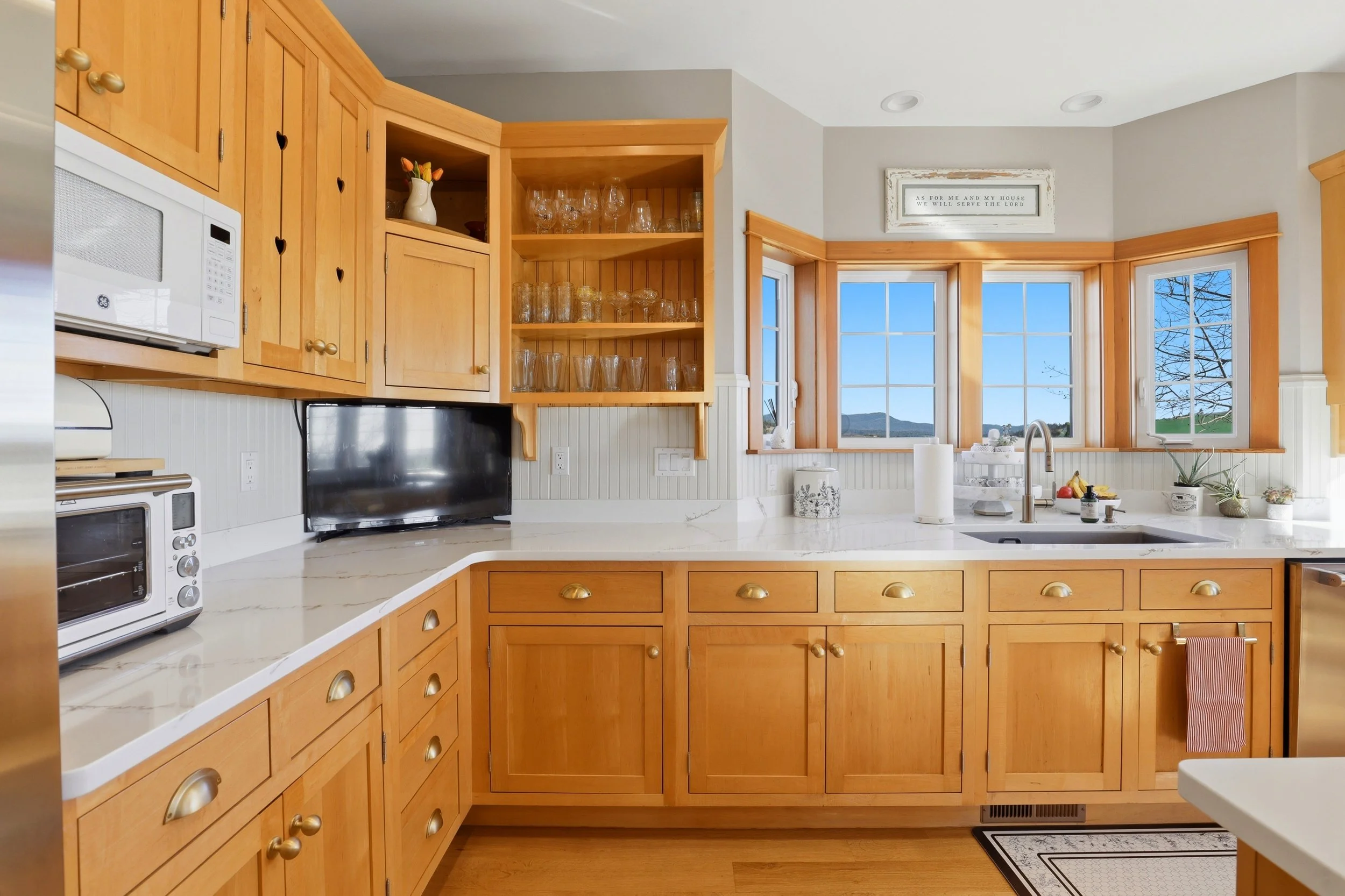 A bright kitchen with natural wood cabinets, white marble countertops, and a large window with mountain and blue sky view. There are glassware in open shelves, a microwave, a TV, and various kitchen decor items.