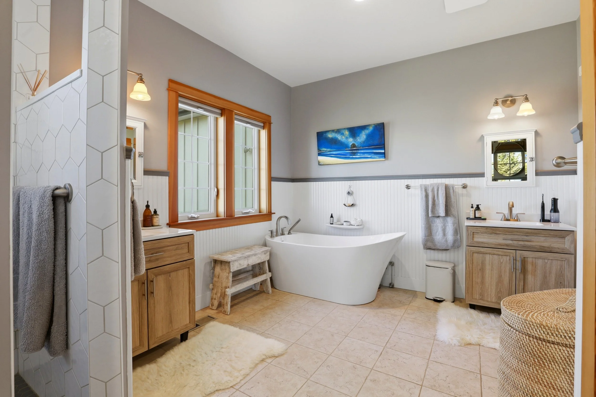 A spacious bathroom with two large windows, a white bathtub, a wooden vanity, and a shower area with hexagonal tile wall. The room features light-colored tile flooring, a fluffy rug, and wall-mounted lighting fixtures.