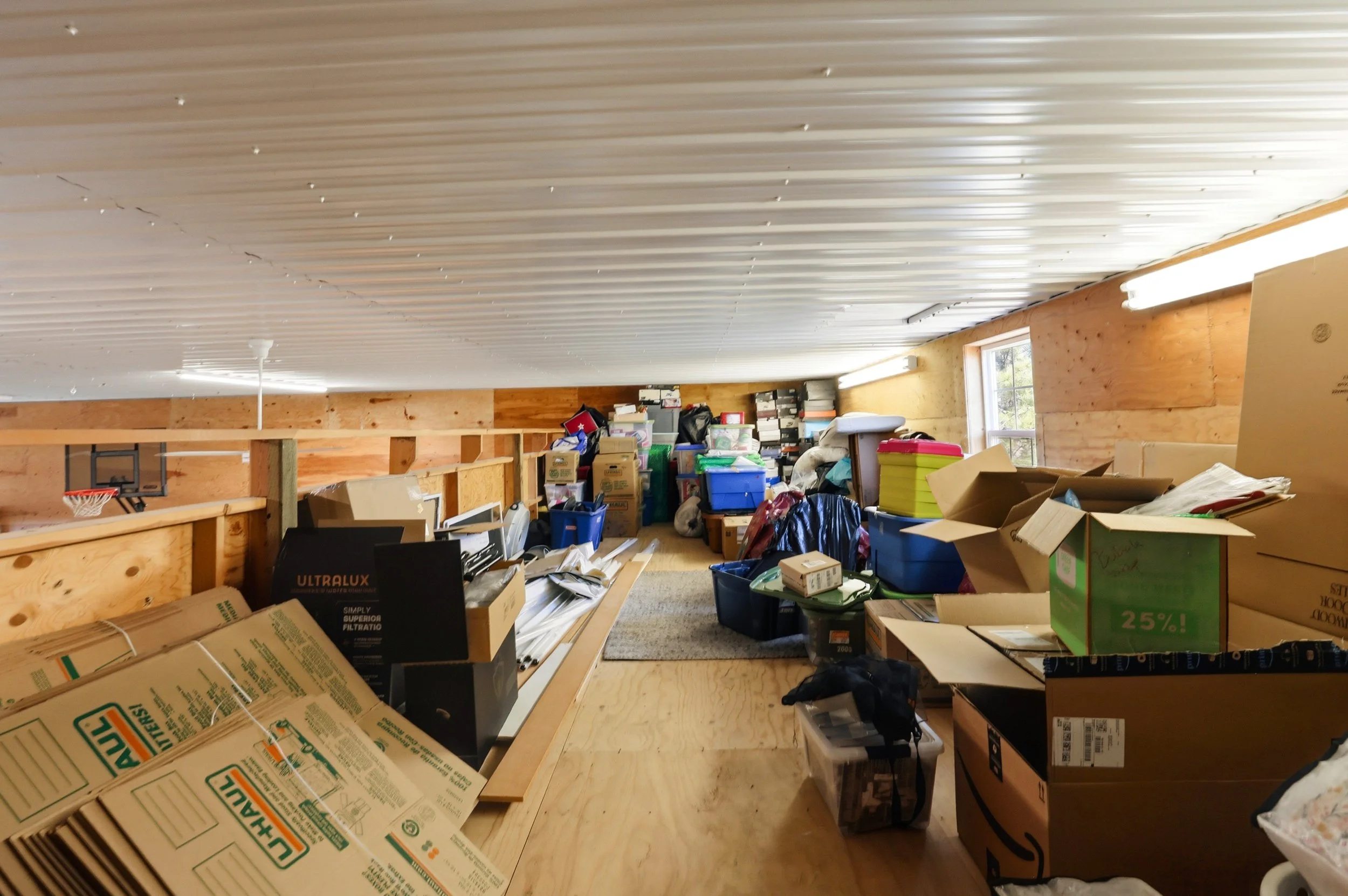 A cluttered room with a sloped white ceiling, wooden walls, a window, and various boxes, papers, and miscellaneous items scattered across the floor and surfaces.