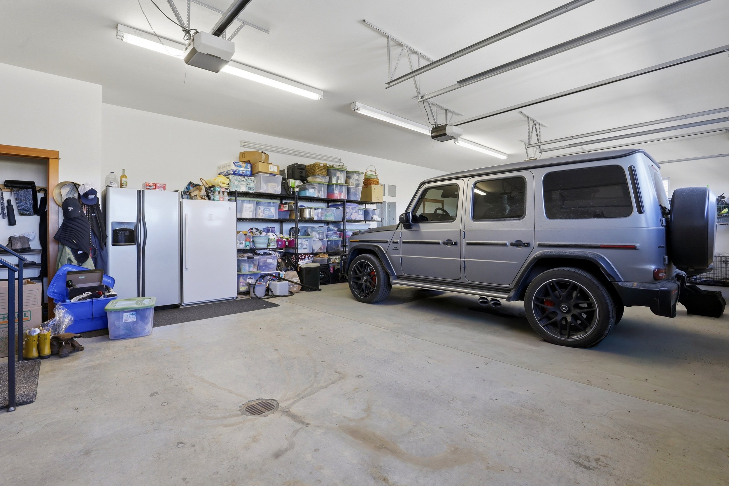 A garage with a gray Mercedes-Benz G-Class SUV parked inside, surrounded by shelves and storage bins, with a refrigerator and various miscellaneous items.