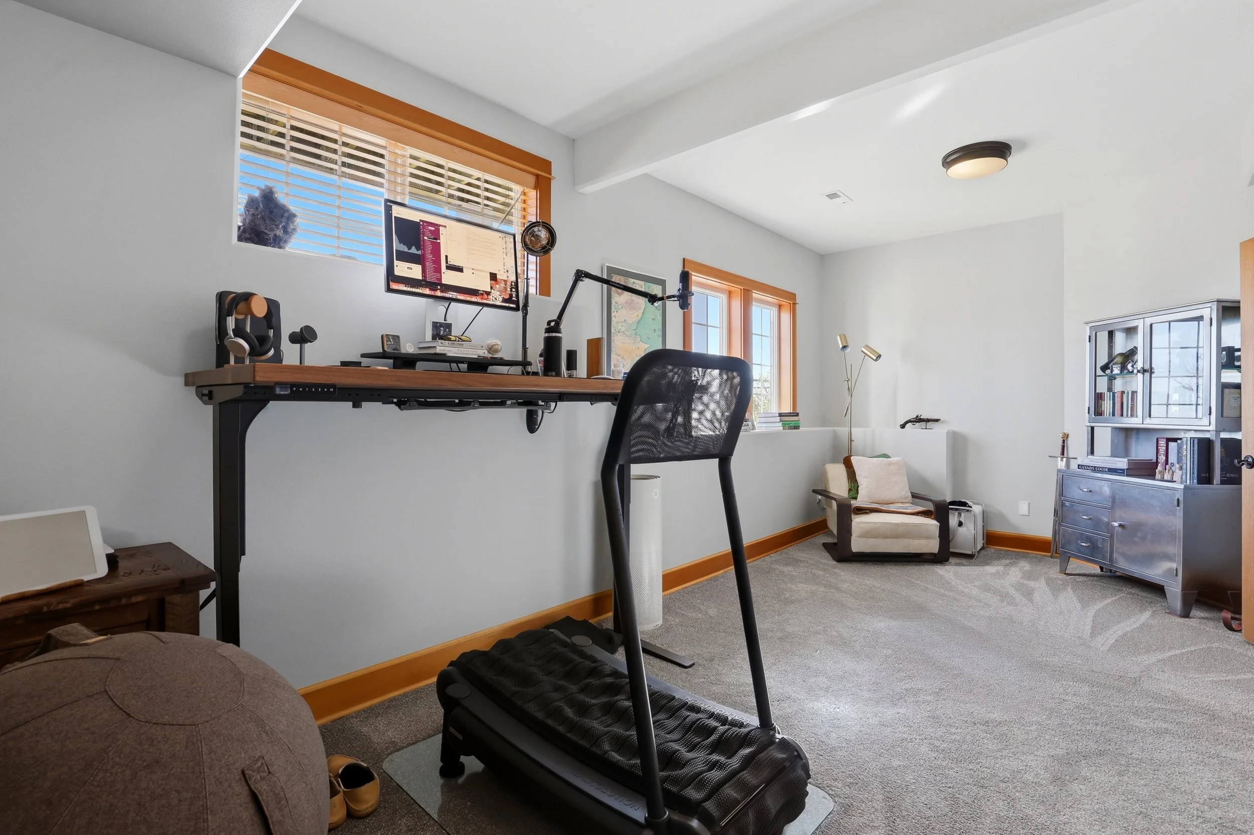 A home office with a wall-mounted desk, a computer monitor, a black office chair, a treadmill, a window with blinds, a bookshelf, and a small seating area.