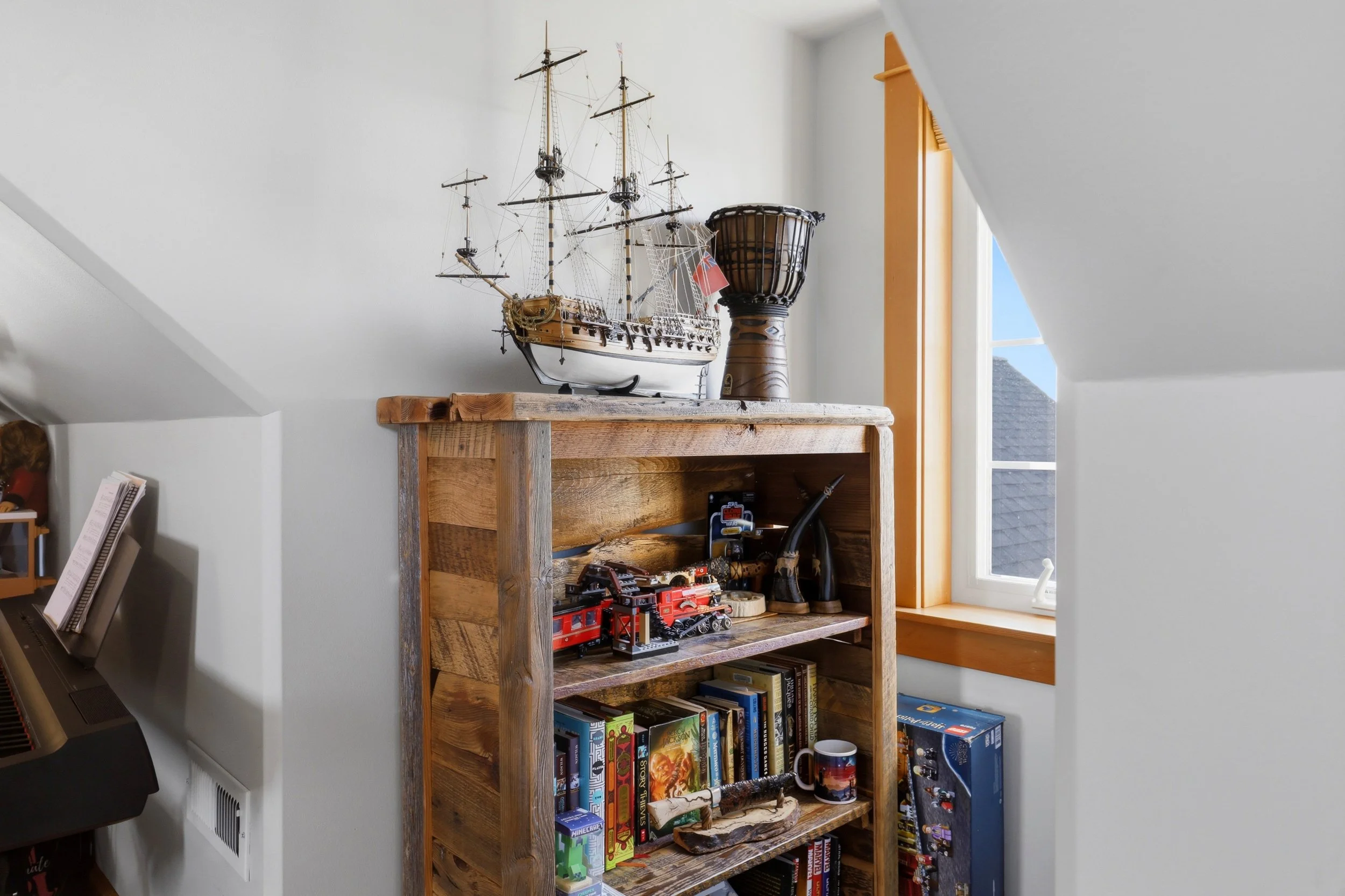 A wooden bookshelf filled with board games and books, a model ship, a large decorative wooden vase, a window with natural light, and a corner of a musical keyboard are visible in a room with white walls and sloped ceiling.