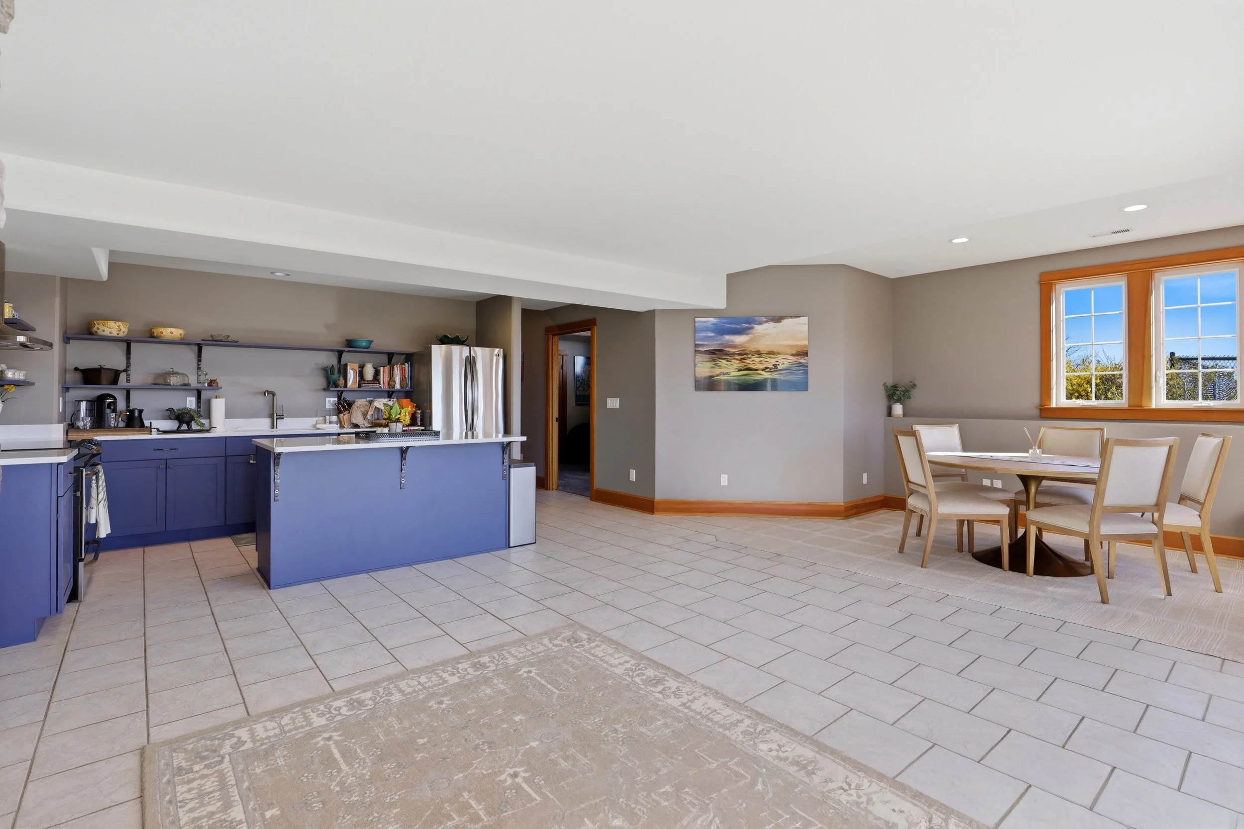 Open-concept kitchen and dining area with beige tiled flooring, blue kitchen cabinets, and a dining table with chairs near large windows showing a view of outside trees.