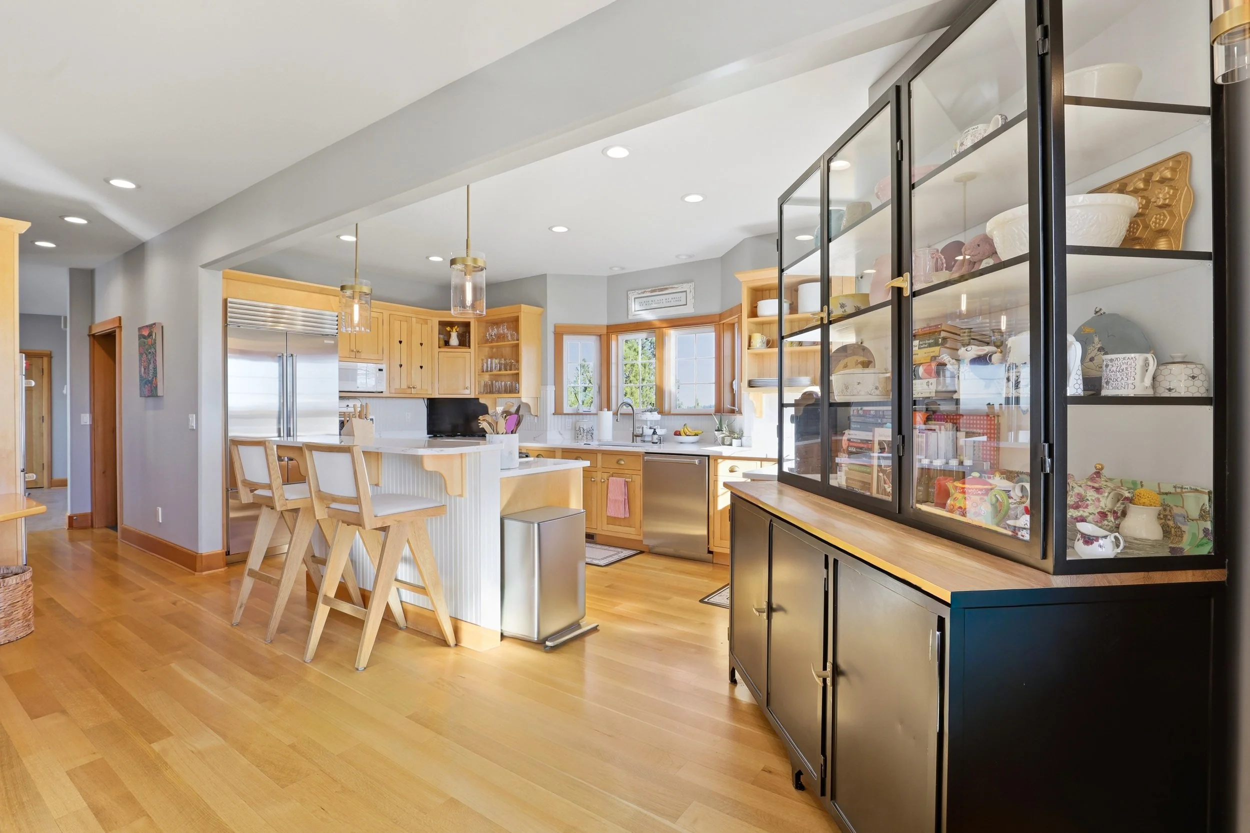 A kitchen with wooden cabinets, a small island with wooden chairs, a stainless steel refrigerator, and a large black cabinet filled with dishes and decorative items, illuminated by recessed ceiling lights and pendant lights.