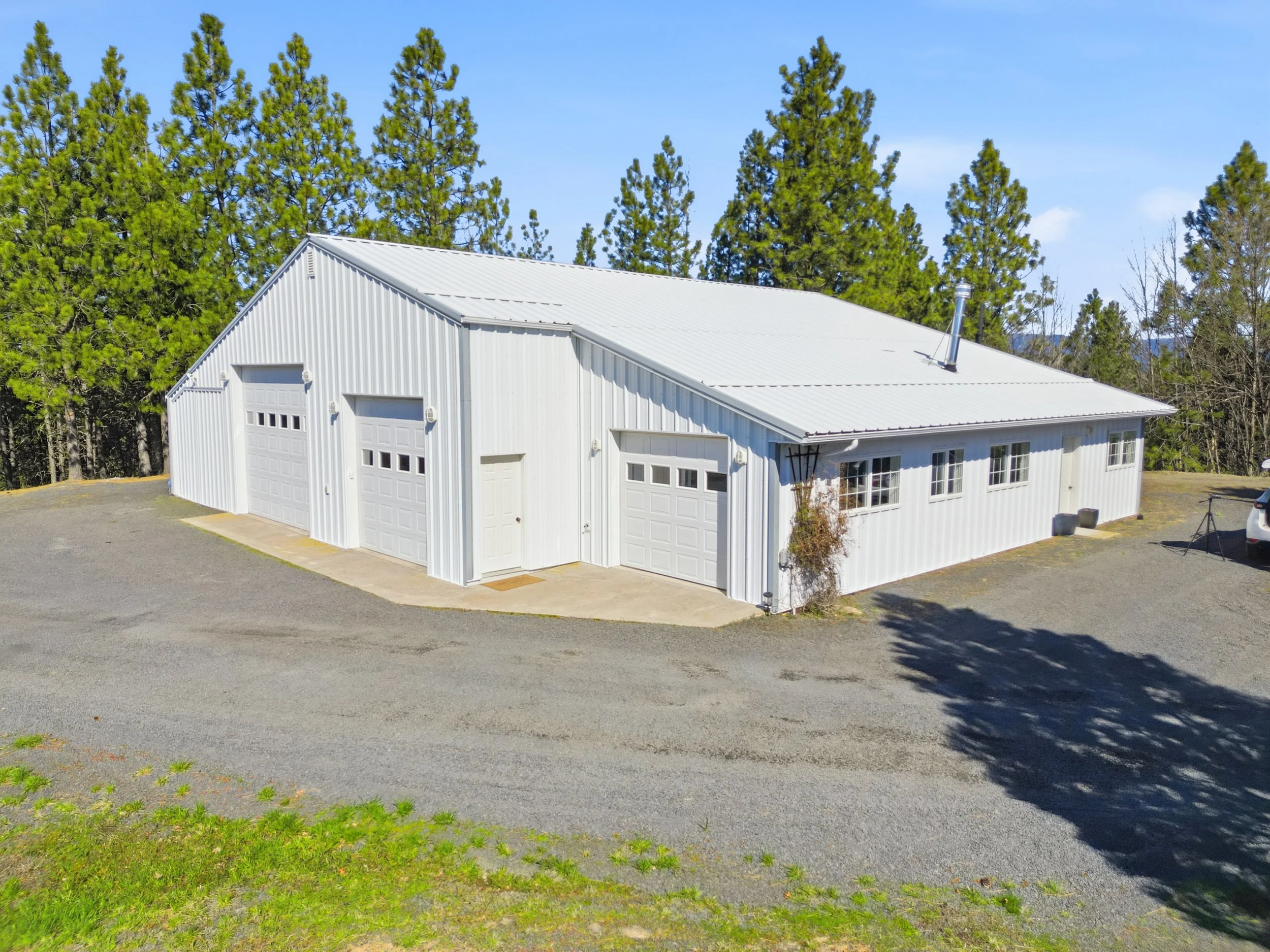 White metal building with multiple garage doors, set against a background of tall green pine trees under a clear blue sky.