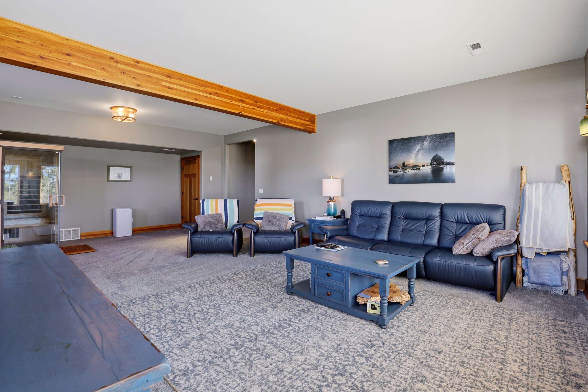 Living room with gray walls, blue leather sofa, matching armchairs, and a wooden coffee table. Decorative pillows, a lamp, sea-themed wall art, window with blinds, and a visible sauna room.