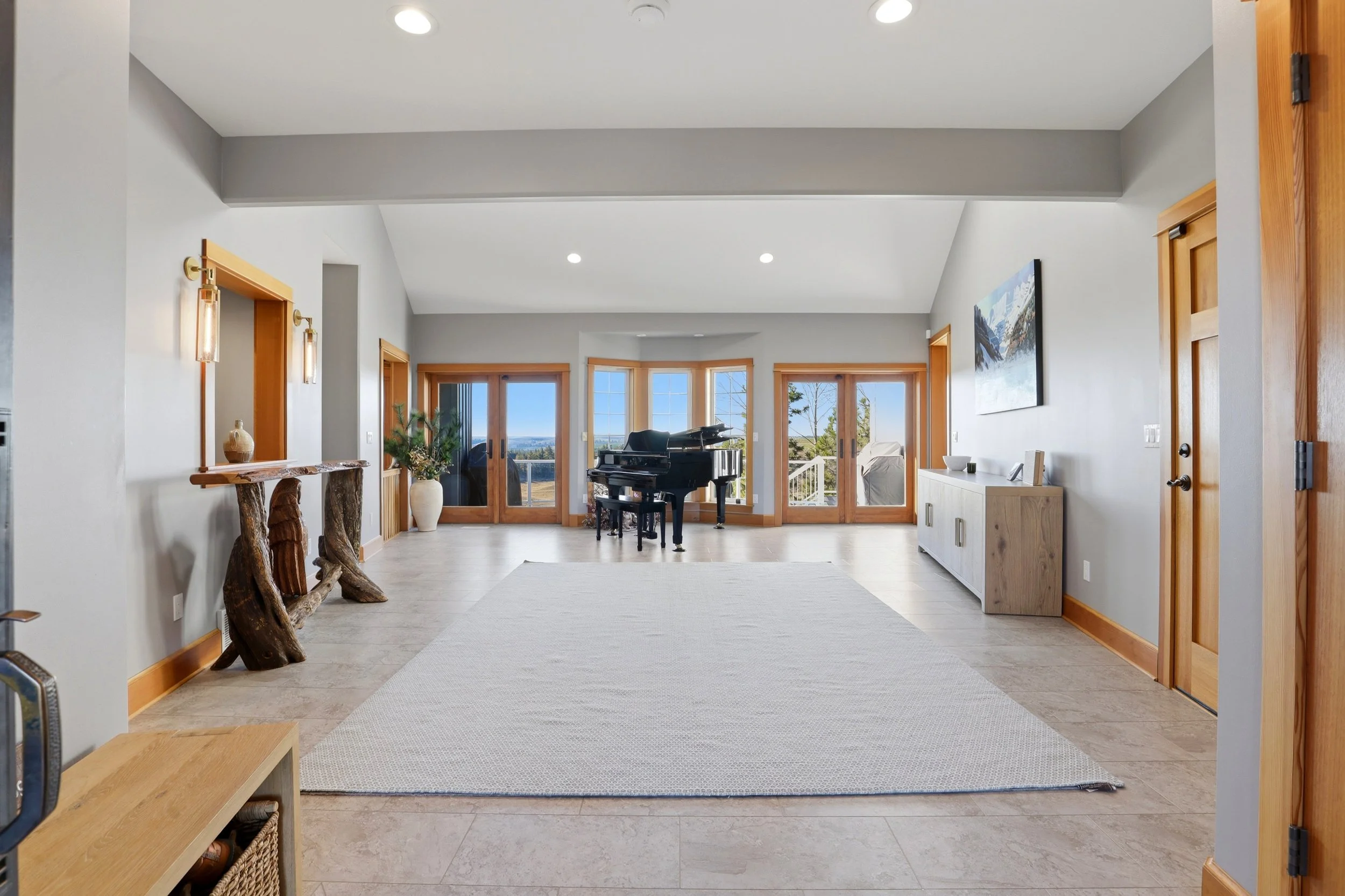 A living room with a grand piano near large glass doors and windows, hardwood trim, and a landscape view outside. There is a textured light-colored rug and wooden accents.
