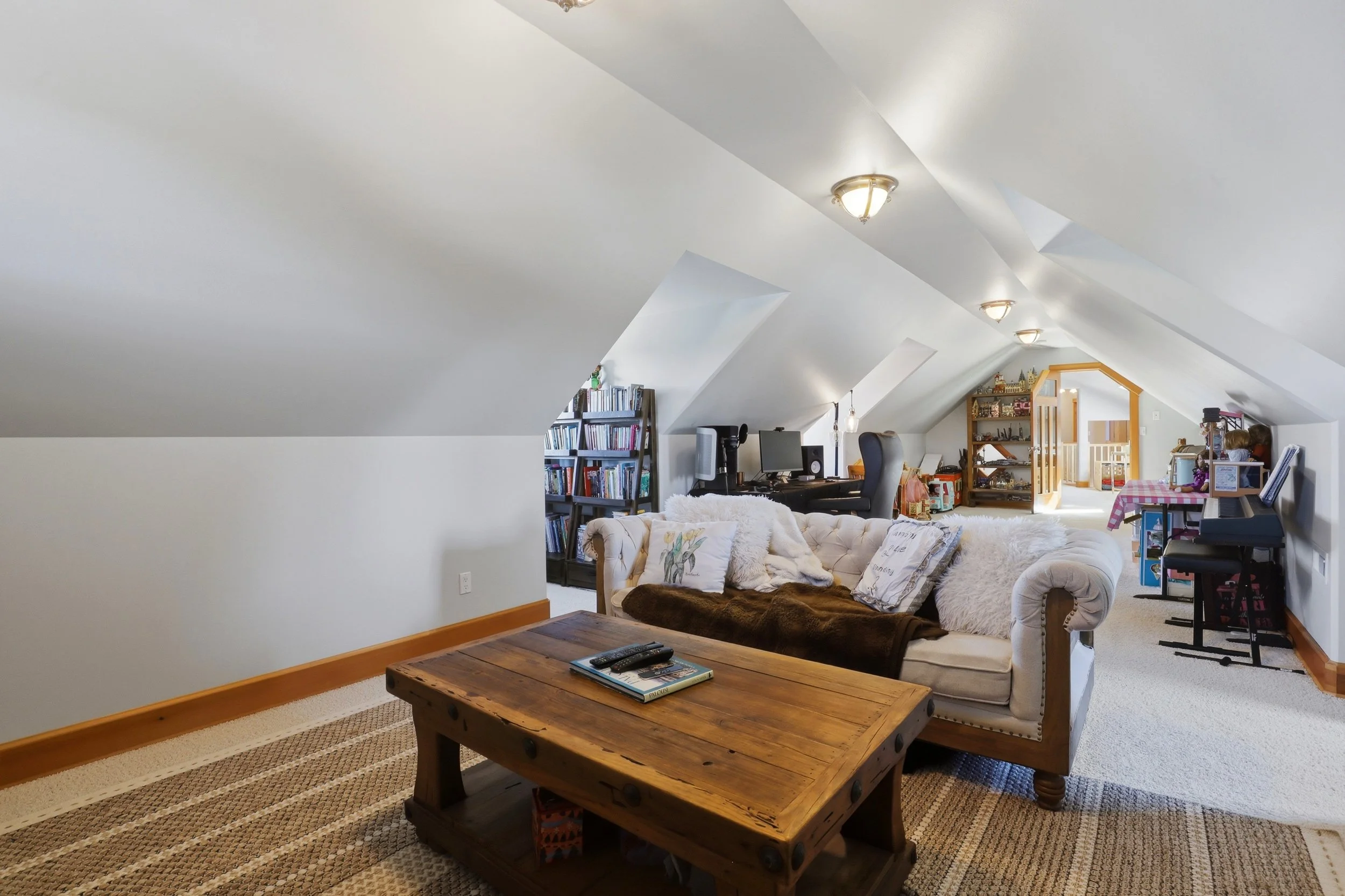 Living room with sloped ceiling, featuring a wooden coffee table, a cream sofa with pillows, a bookshelf, a desk with a computer, and various decorative items, illuminated by ceiling lights.