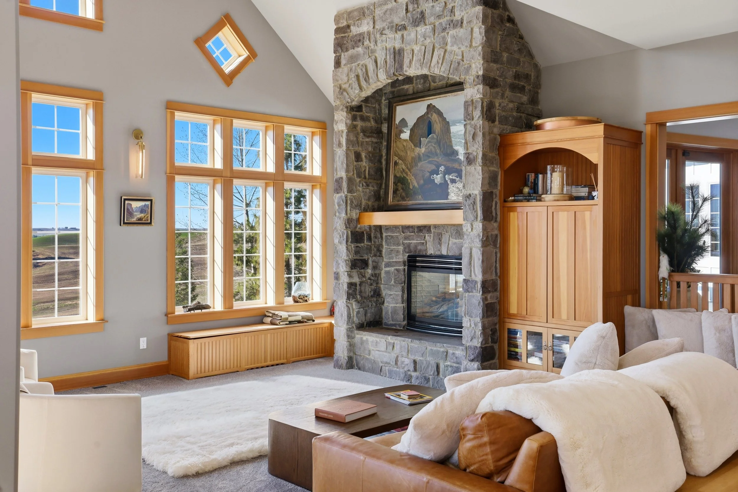 A living room featuring large windows with wooden frames, a stone fireplace with a painting above it, wooden cabinetry, a beige sofa with pillows, and a cream-colored armchair, all with a view of a bright, outdoor landscape.