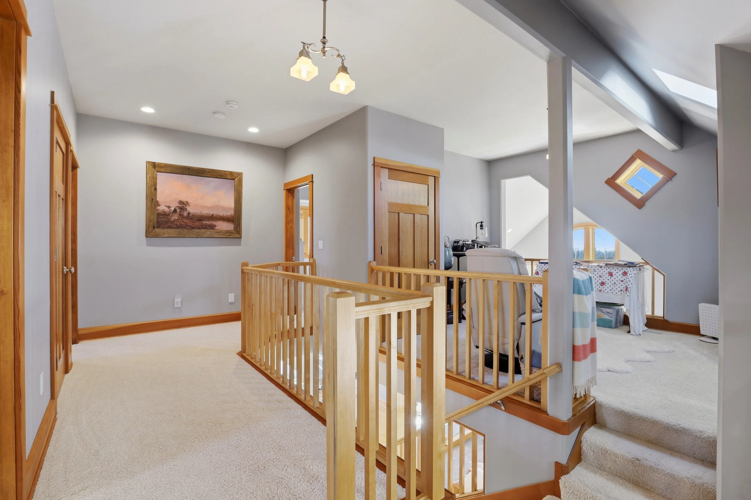 Interior view of an upper landing area with beige carpeting, wooden railing, walls painted light gray, artwork on the wall, open doorways, and a bathroom visible in the background. There are ceiling lights and a ceiling-mounted chandelier with two gl