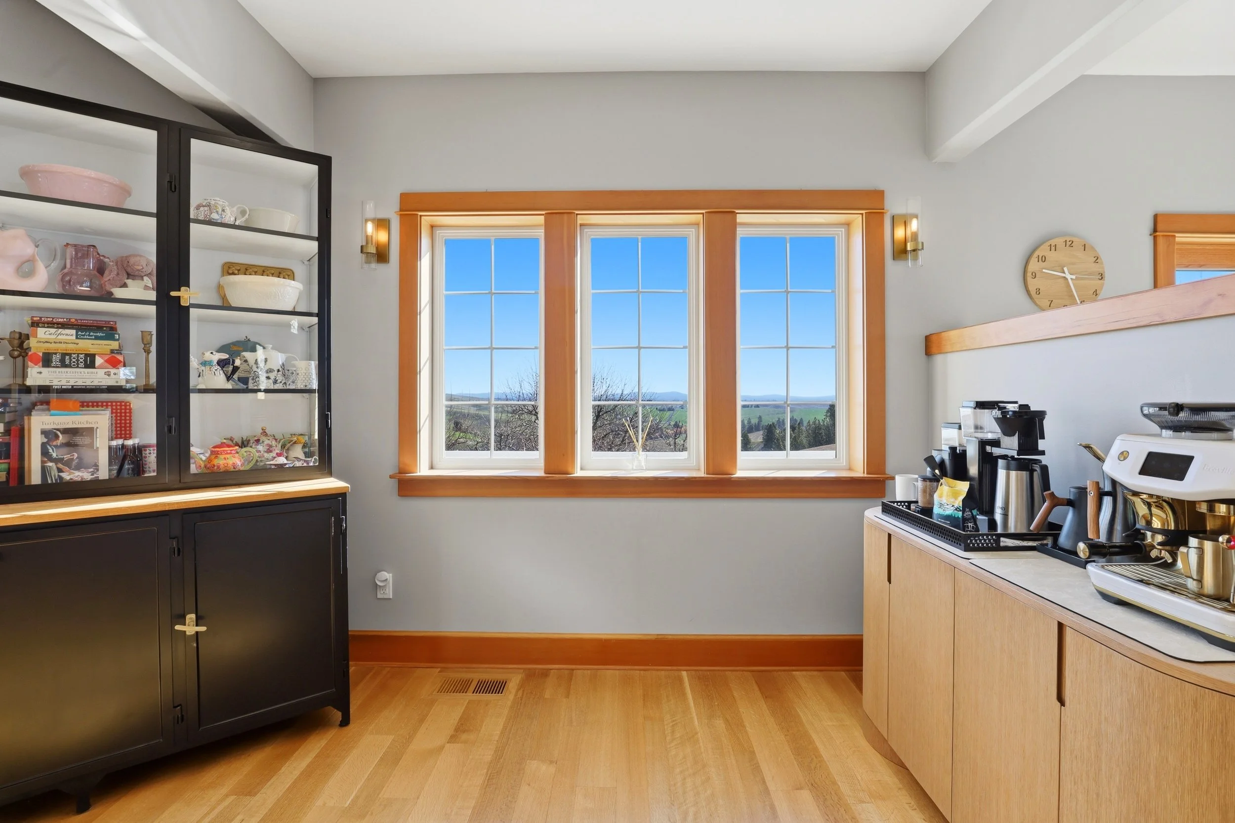 A kitchen with a large window showing a scenic outdoors view, black china cabinet on the left, wooden cabinets and countertop on the right, coffee machine and kitchen utensils on the counter, and a clock on the wall.
